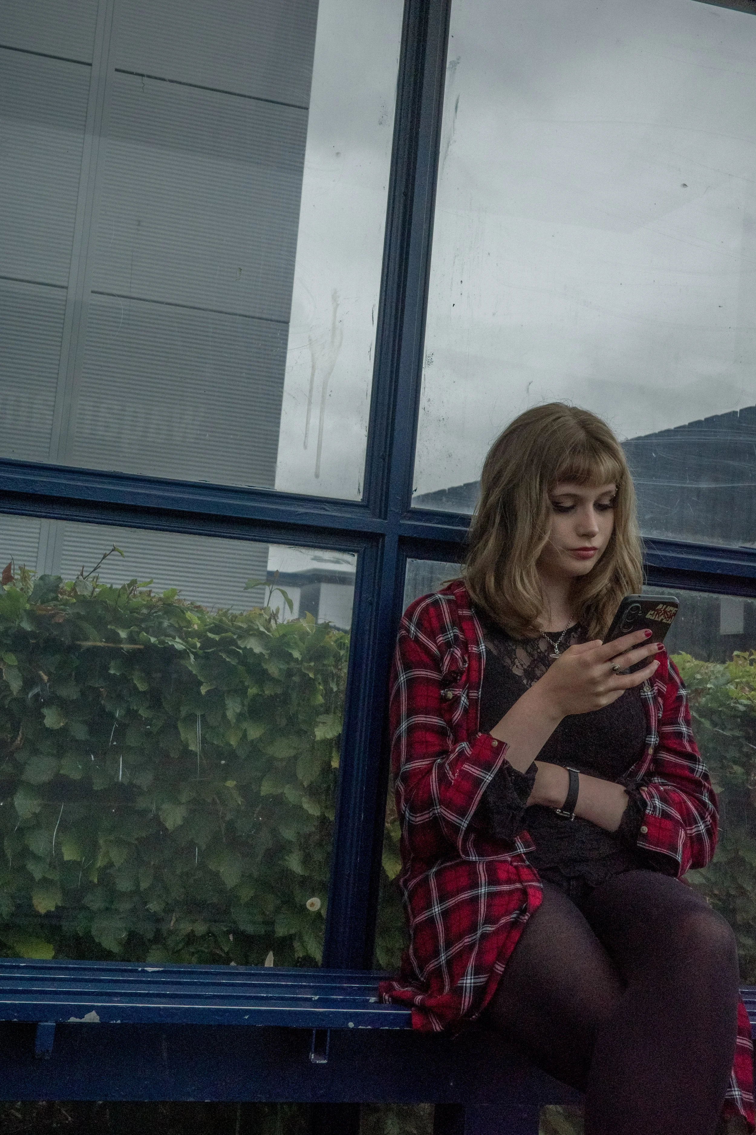 A young woman with blonde hair wearing a red plaid shirt and black lace top, sitting on a blue bench and looking at her phone. Behind her, there are green bushes and a large window with a metal frame.