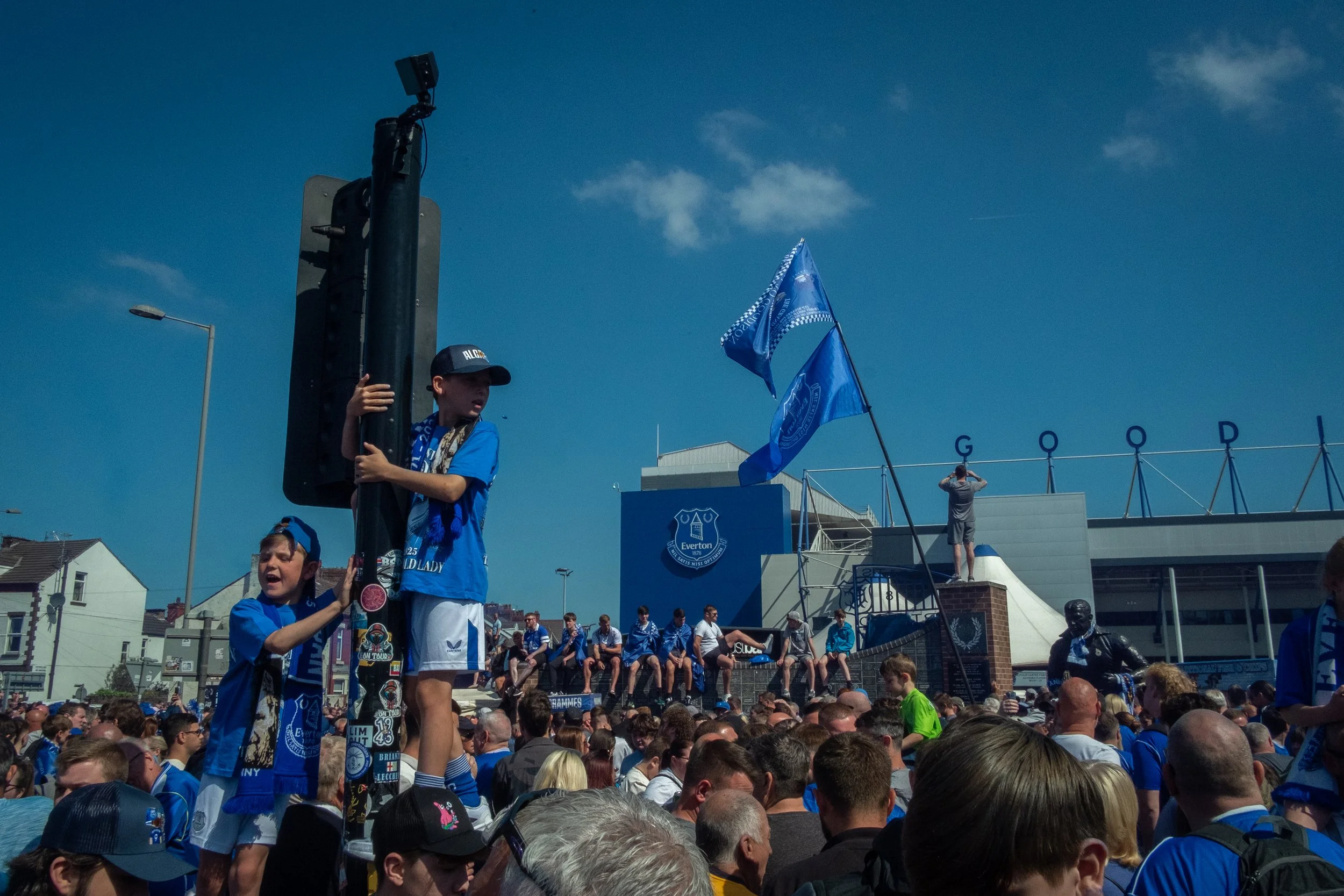 Crowd of people at an outdoor football stadium, with two kids in blue sports shirts and scarves standing on a platform, one holding a pole with a flag, in front of the Everton Football Club stadium.