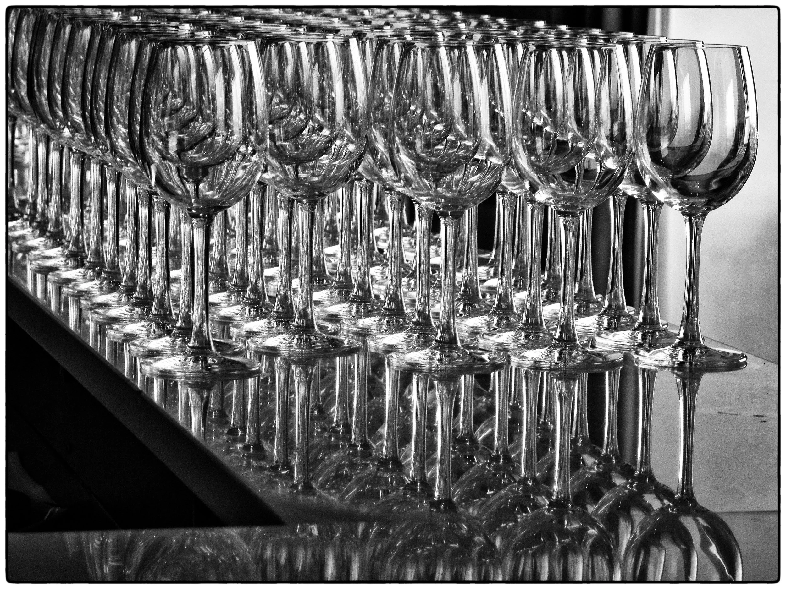 Multiple empty wine glasses arranged in rows on a table, reflected on the surface below in black and white.