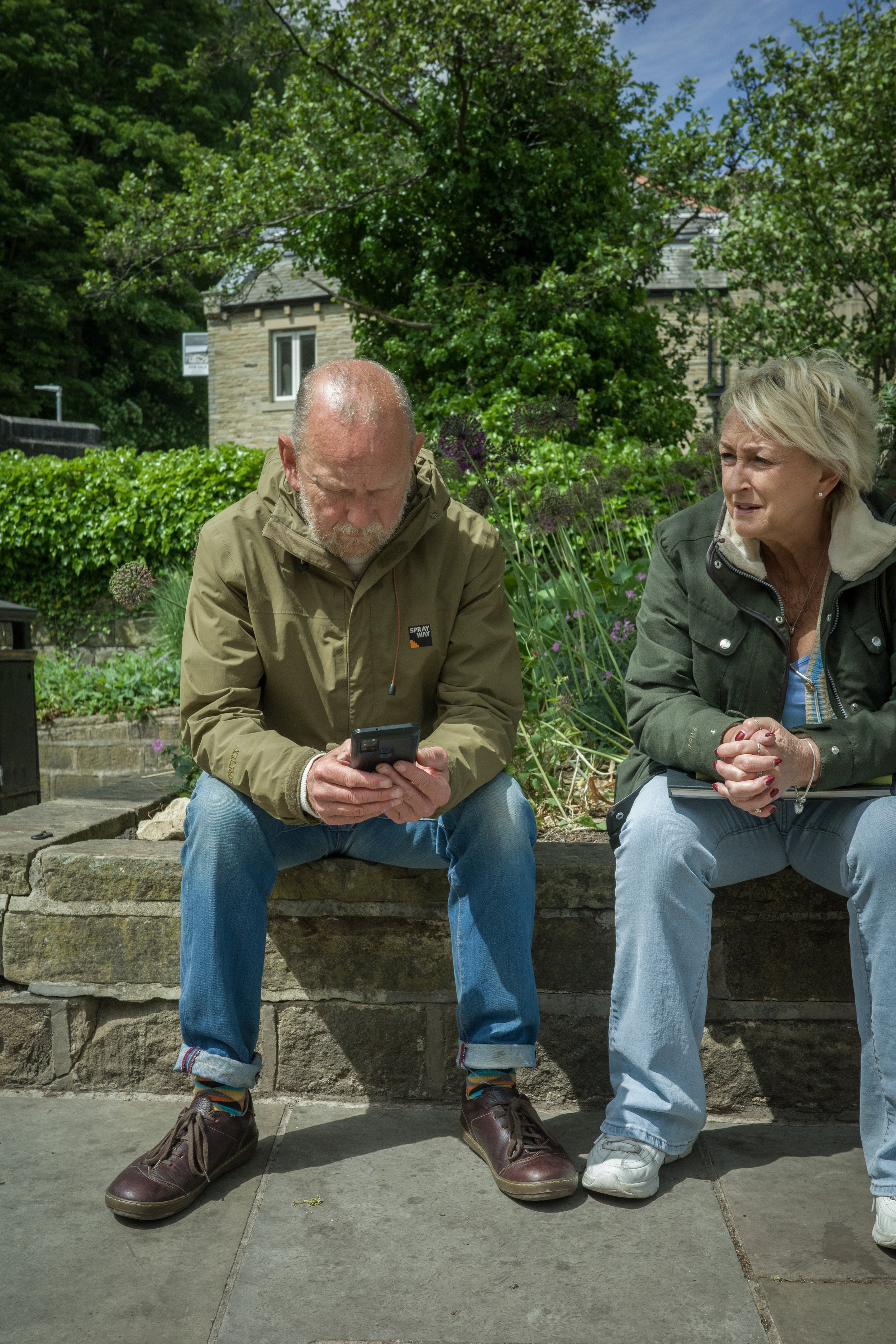 An older man with a beard wearing a khaki jacket and blue jeans sitting on a stone bench, looking at his phone, beside an older woman with short blonde hair wearing a green jacket and light blue jeans, sitting on the same bench, with a garden and a h