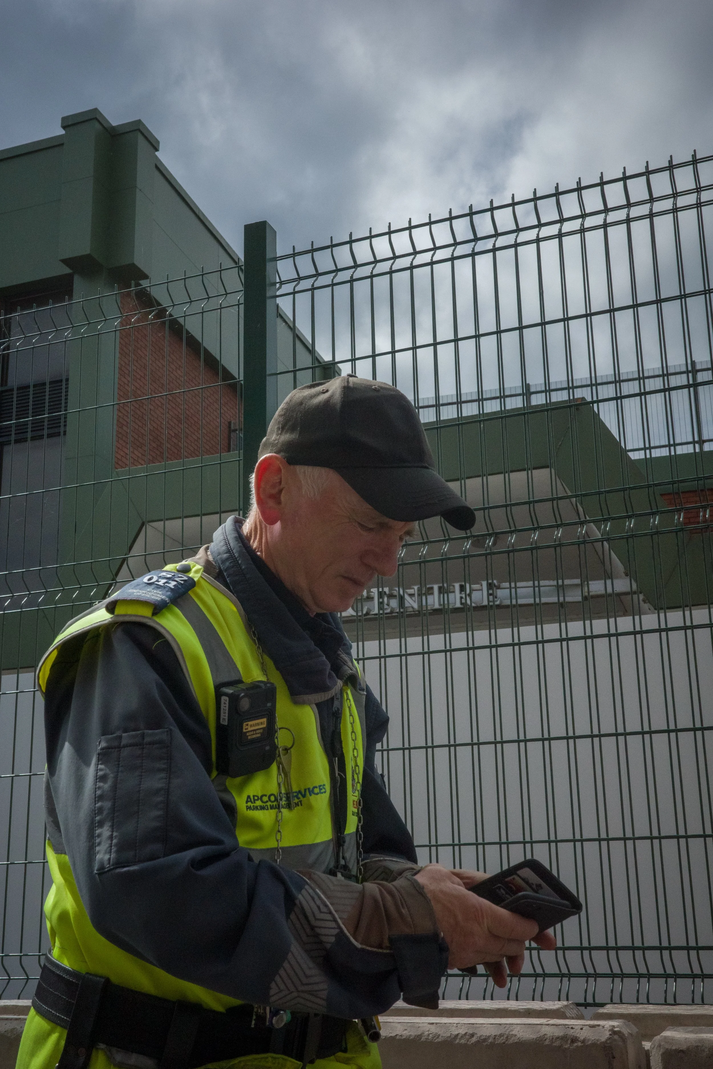 A man in a grey and yellow high-visibility jacket with a headset and gloves, holding a tablet, standing in front of a metal fence with a building and cloudy sky behind him.