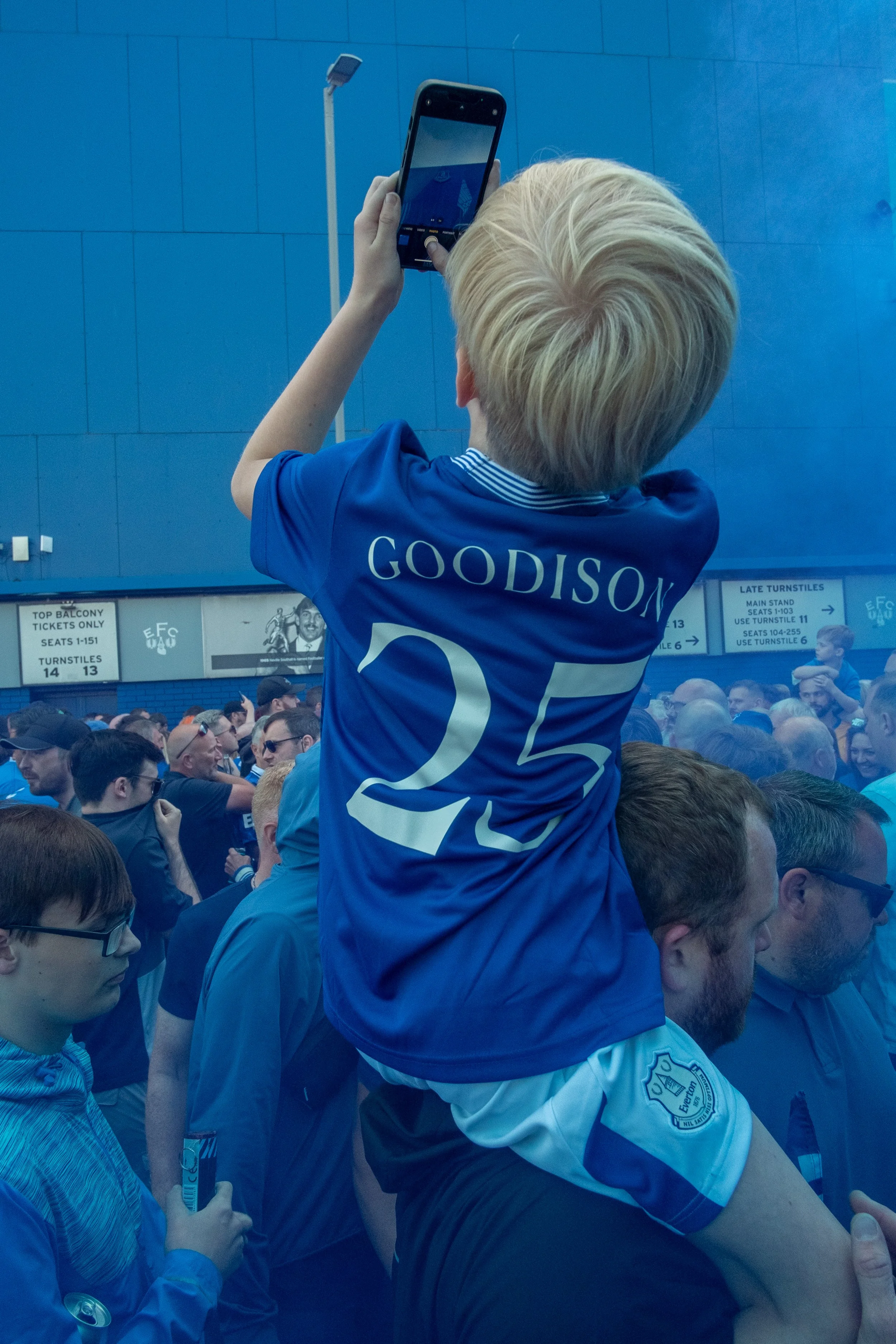 A young boy wearing a blue soccer jersey with the name 'Goodison' and the number 25, sitting on someone's shoulders, taking a photo with a smartphone. The crowd in front appears to be at a sports event, with blue colors and spectators visible.