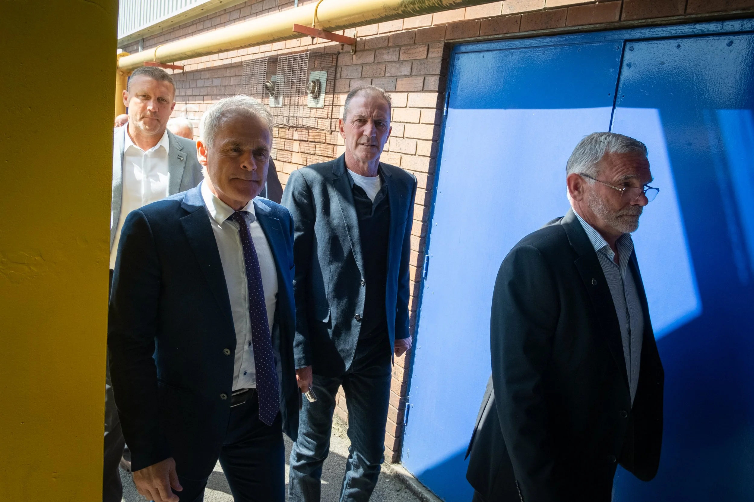Four men in formal suits walking outside near brick walls, one with glasses, with a blue door and a yellow wall in the foreground.