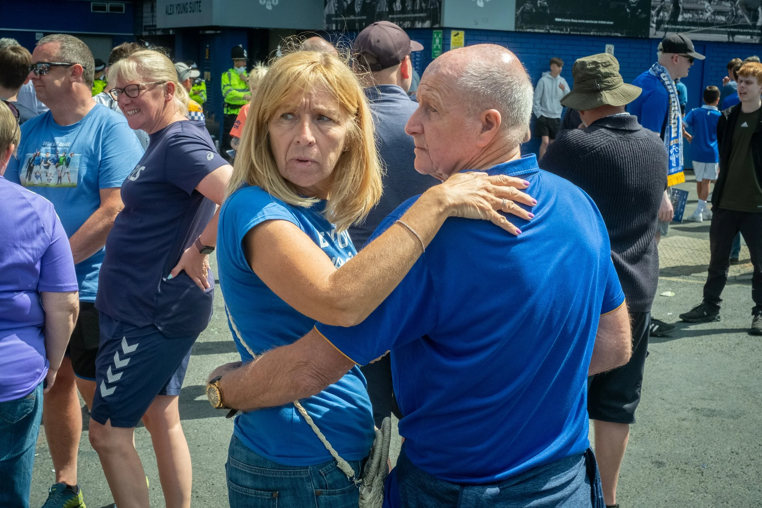 A woman with blonde hair and a man with a bald head sharing an emotional moment outside, surrounded by a crowd of people, some wearing blue shirts, in a stadium or sports venue.