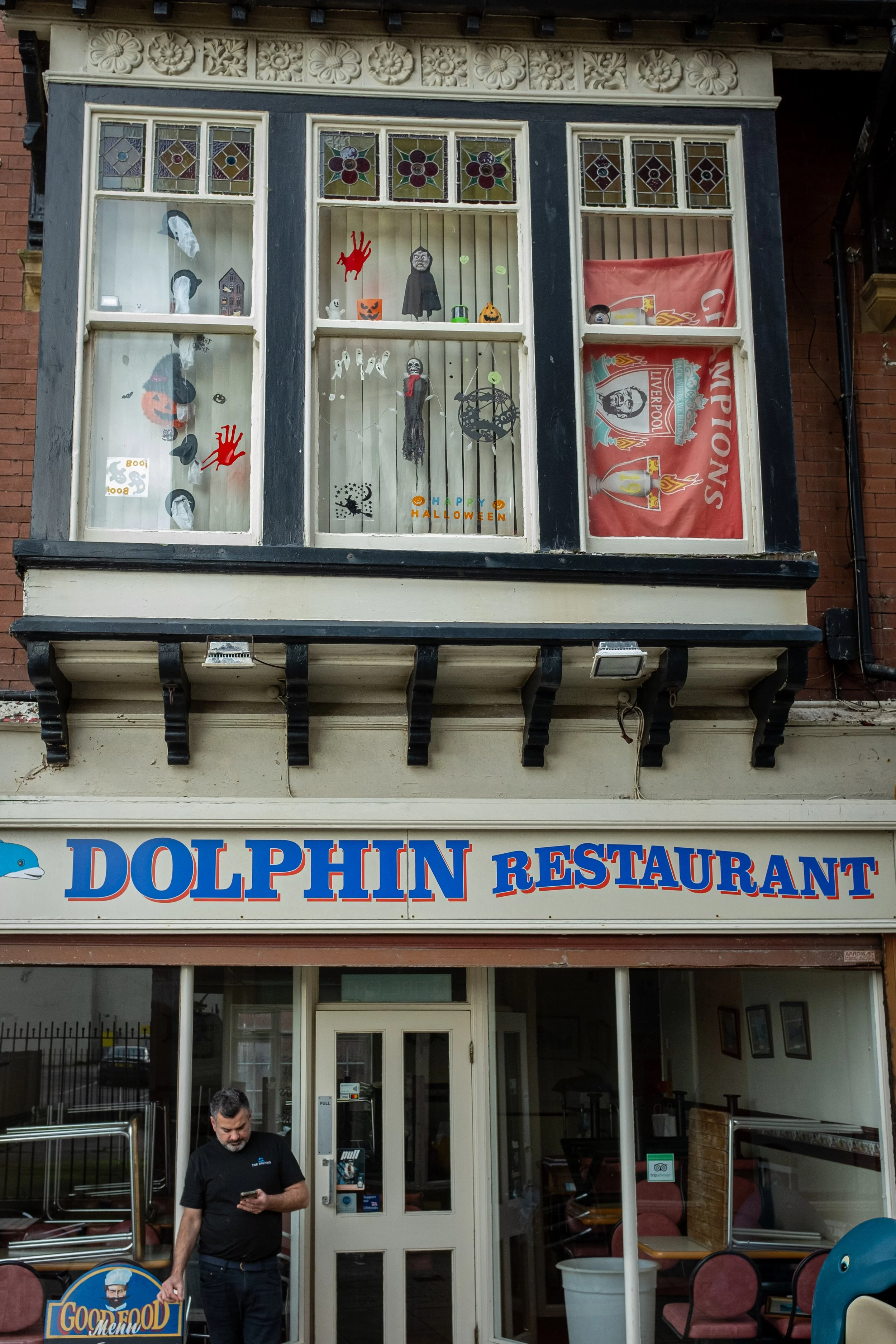 The entrance of the Dolphin Restaurant with a window display decorated for Halloween, featuring Halloween-themed decorations like ghosts, pumpkins, and a skeleton. A man is standing at the entrance looking at his phone, and the restaurant sign is vis