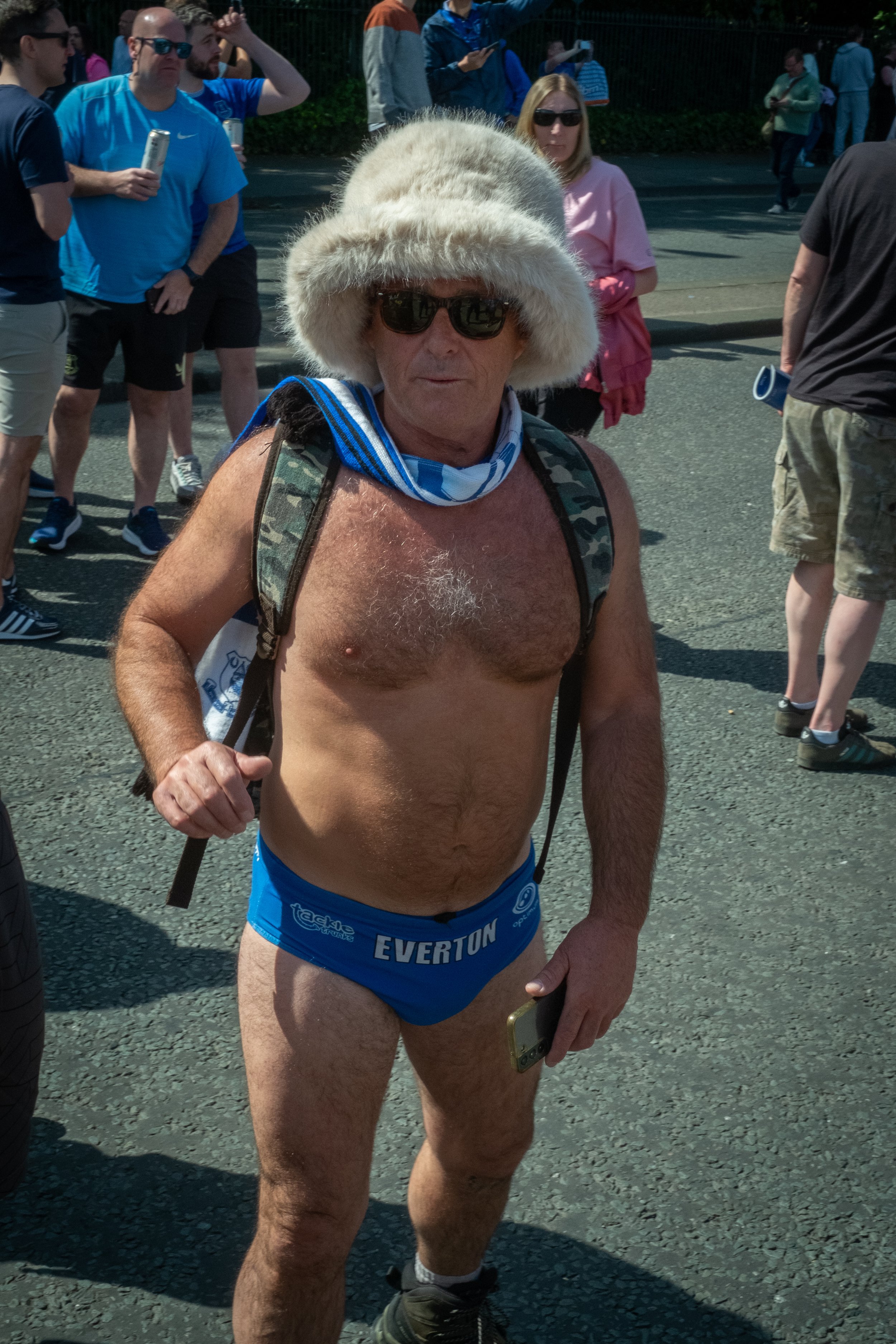 A shirtless man wearing a furry hat, sunglasses, blue running shorts, and a backpack, standing outdoors among a crowd of people.