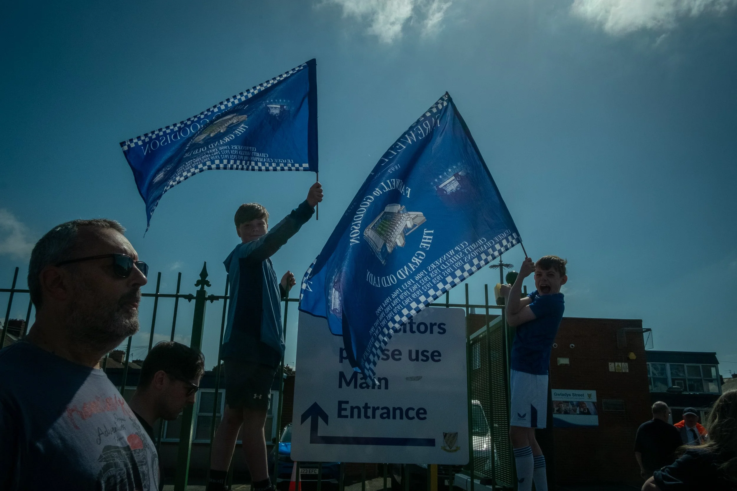 Two boys waving blue flags with a man and other people standing nearby, behind a sign. The scene is outdoors during the daytime with a cloudy sky.