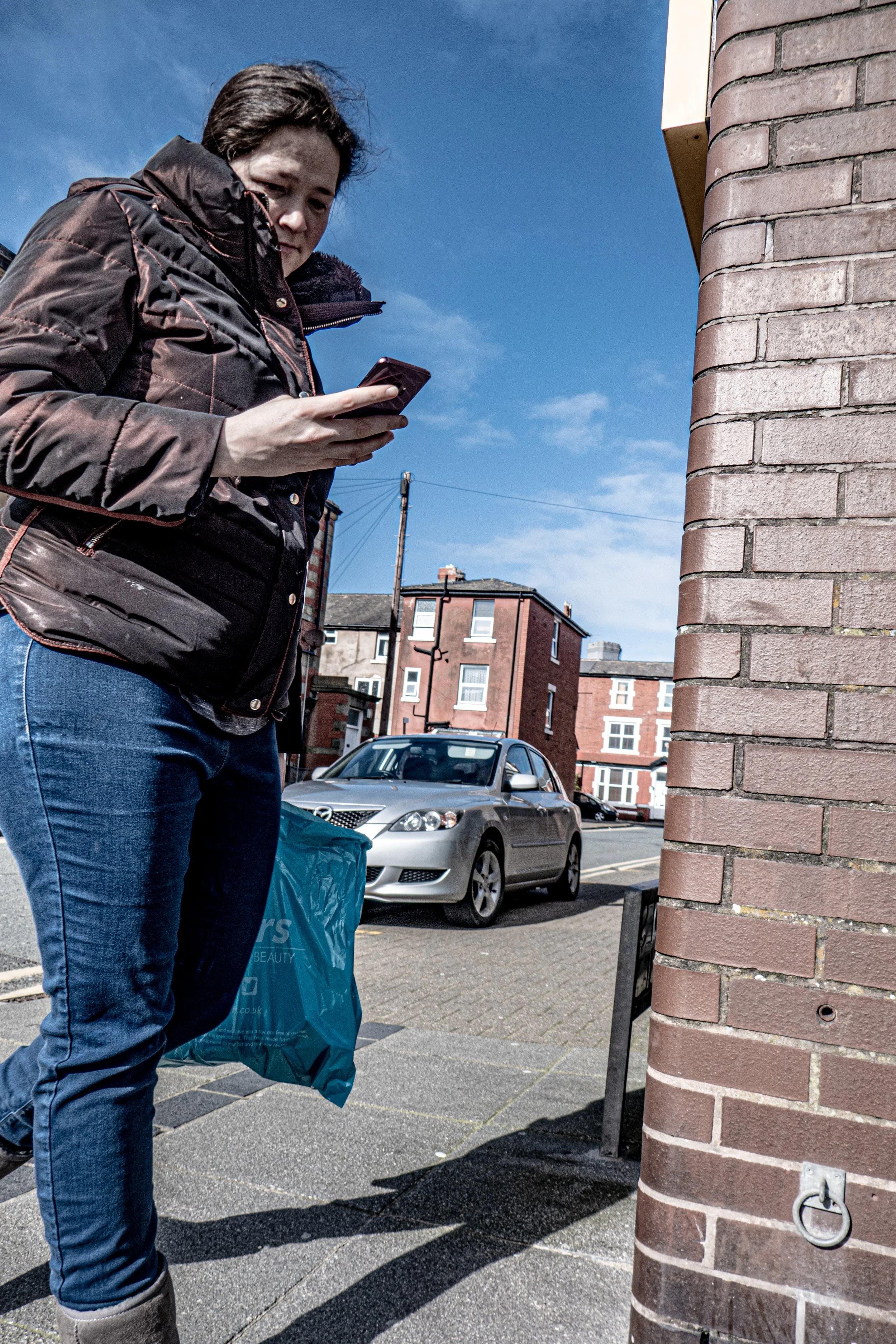 A woman wearing a brown jacket and blue jeans standing near a brick wall, looking at her phone, with cars and buildings in the background under a blue sky.