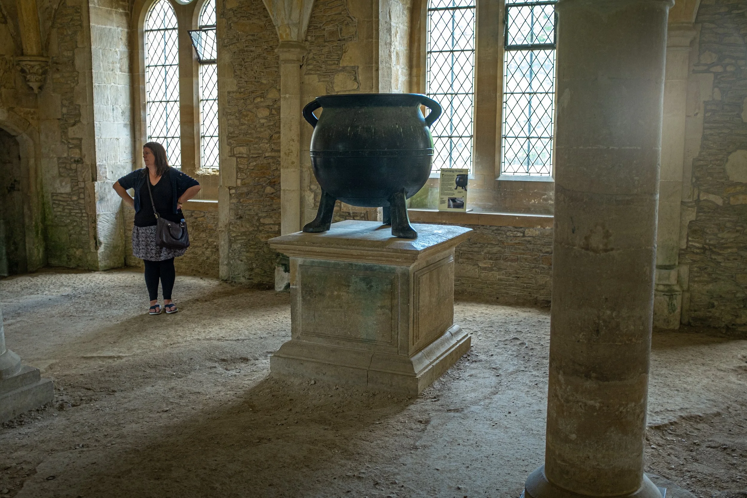 A woman standing inside a historic stone building, looking at a large black ancient urn or cauldron displayed on a pedestal, with tall stone columns and windows in the background.