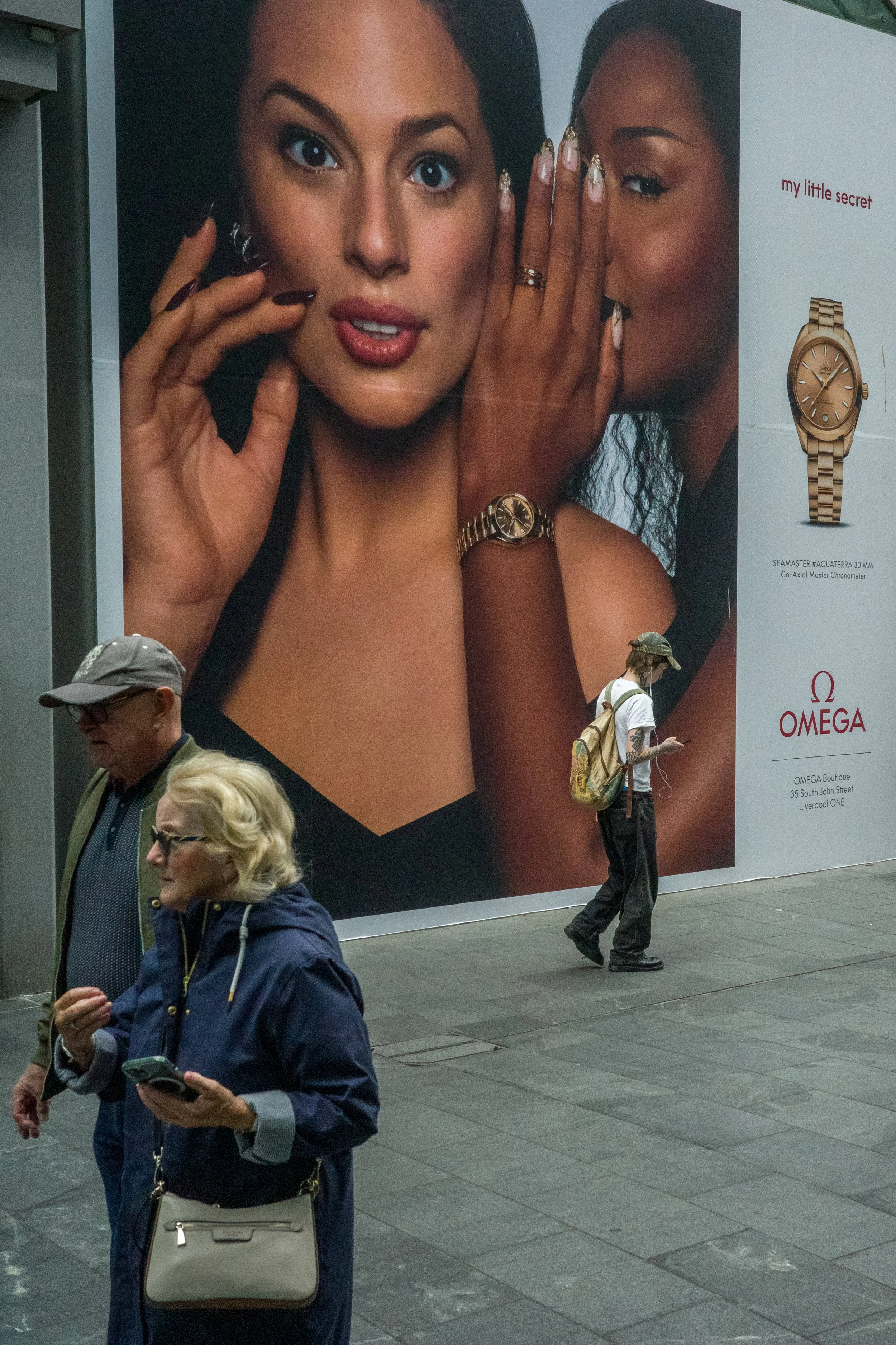 A large advertisement for Omega watches. It features two women, one with dark hair and the other with curly hair, showing off their watches and jewelry. Below, there are several pedestrians walking along the sidewalk, including a woman with blond hai