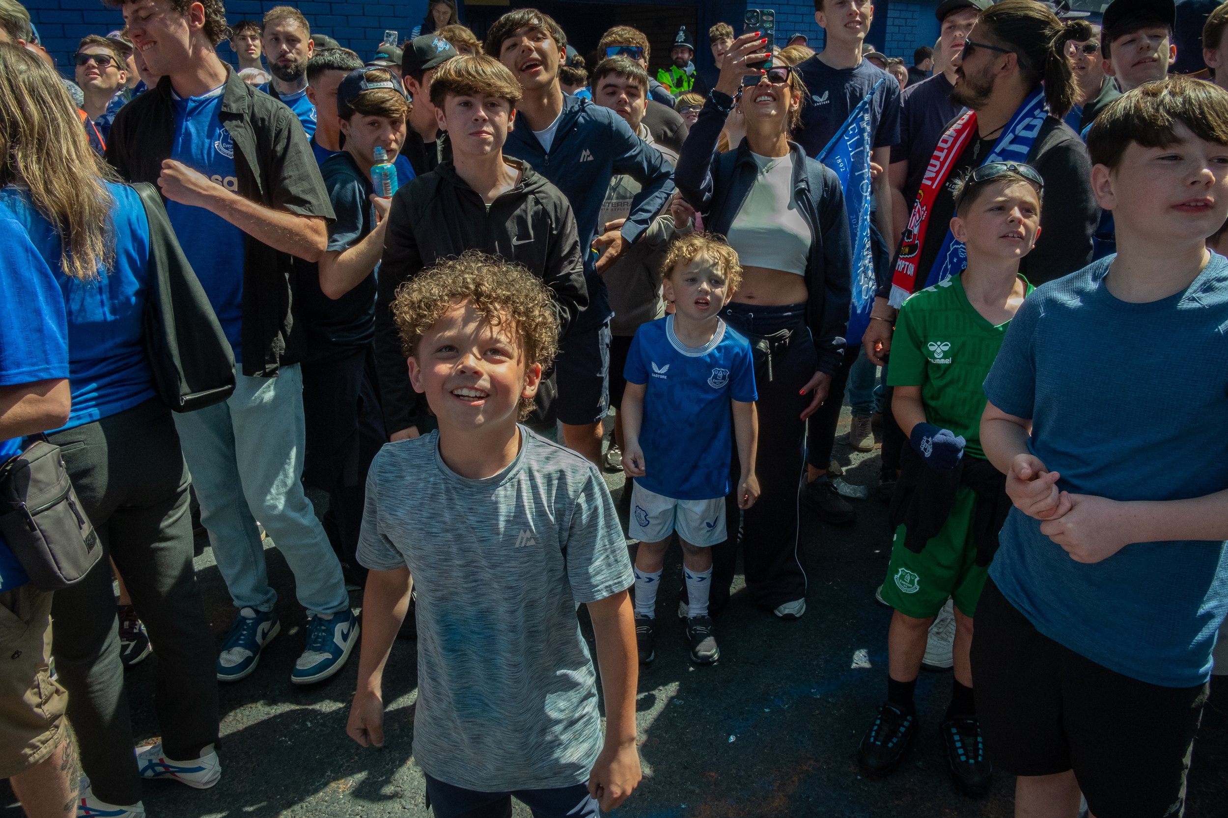 A group of children and adults gathered outdoors, some wearing sports jerseys and scarves, with many smiling and taking photos.
