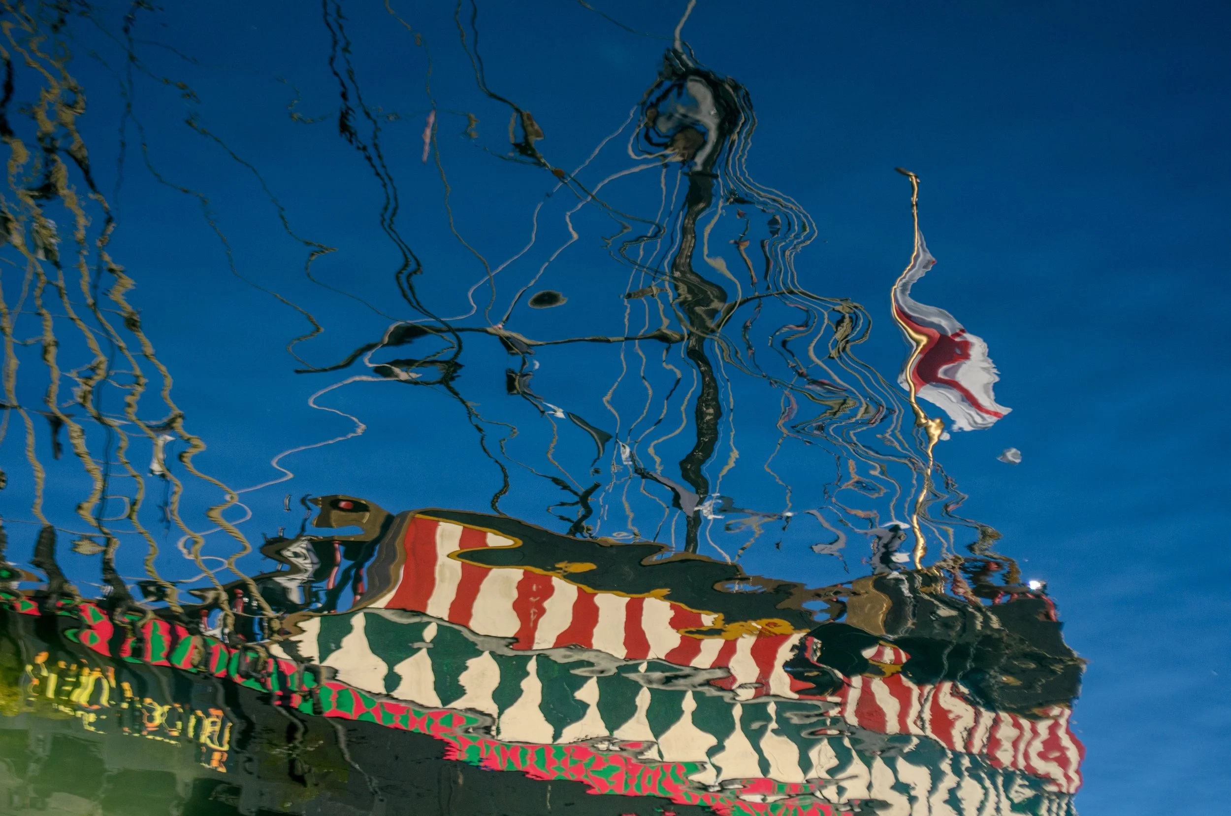 Reflected image of a boat with colorful red, white, green, and black patterns on the water surface with distorted lines of reflections from the boat and nearby structures.