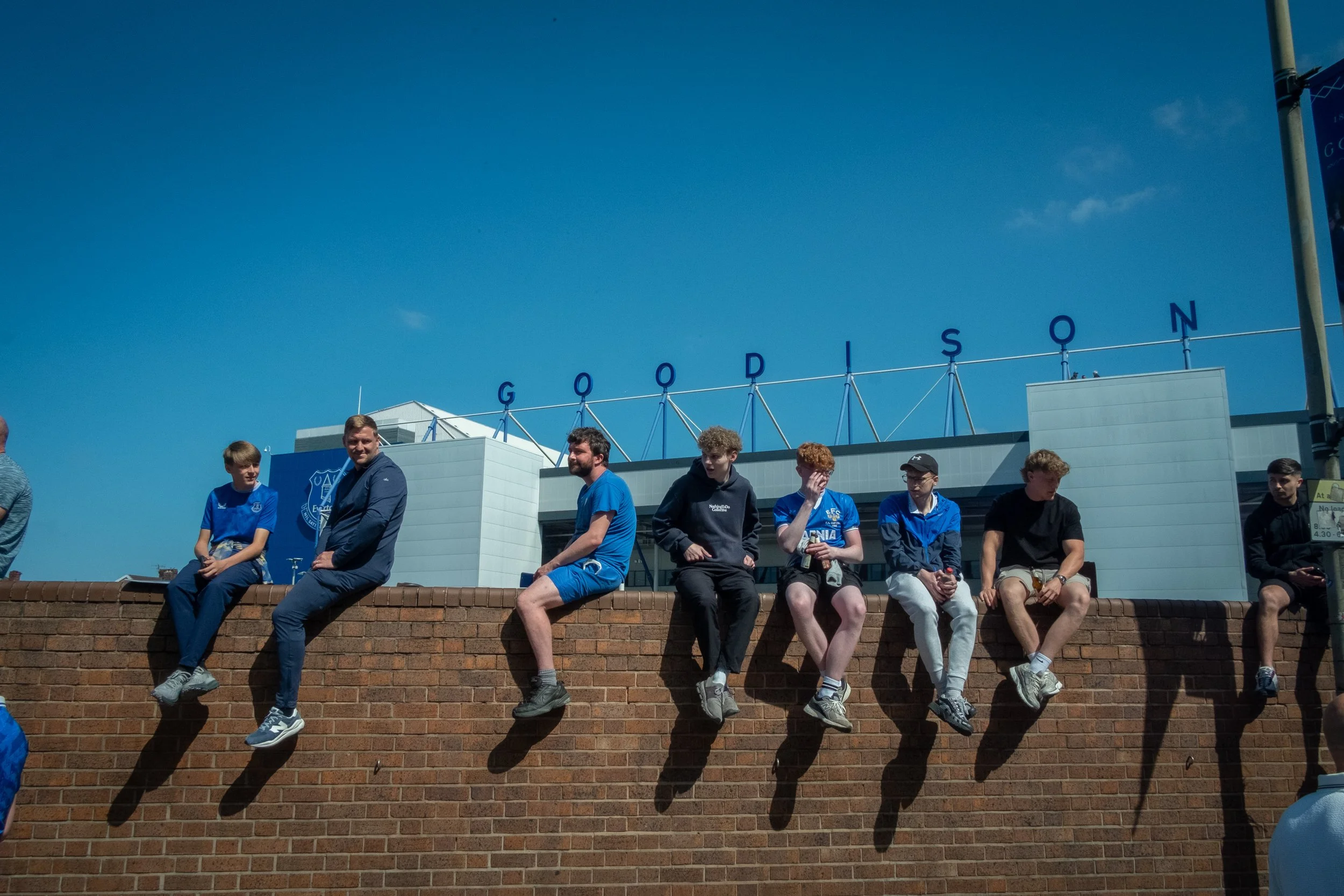 Group of young men sitting on a brick wall outside Everton Football Club stadium on a sunny day, with the 'Goodison' sign visible in the background.