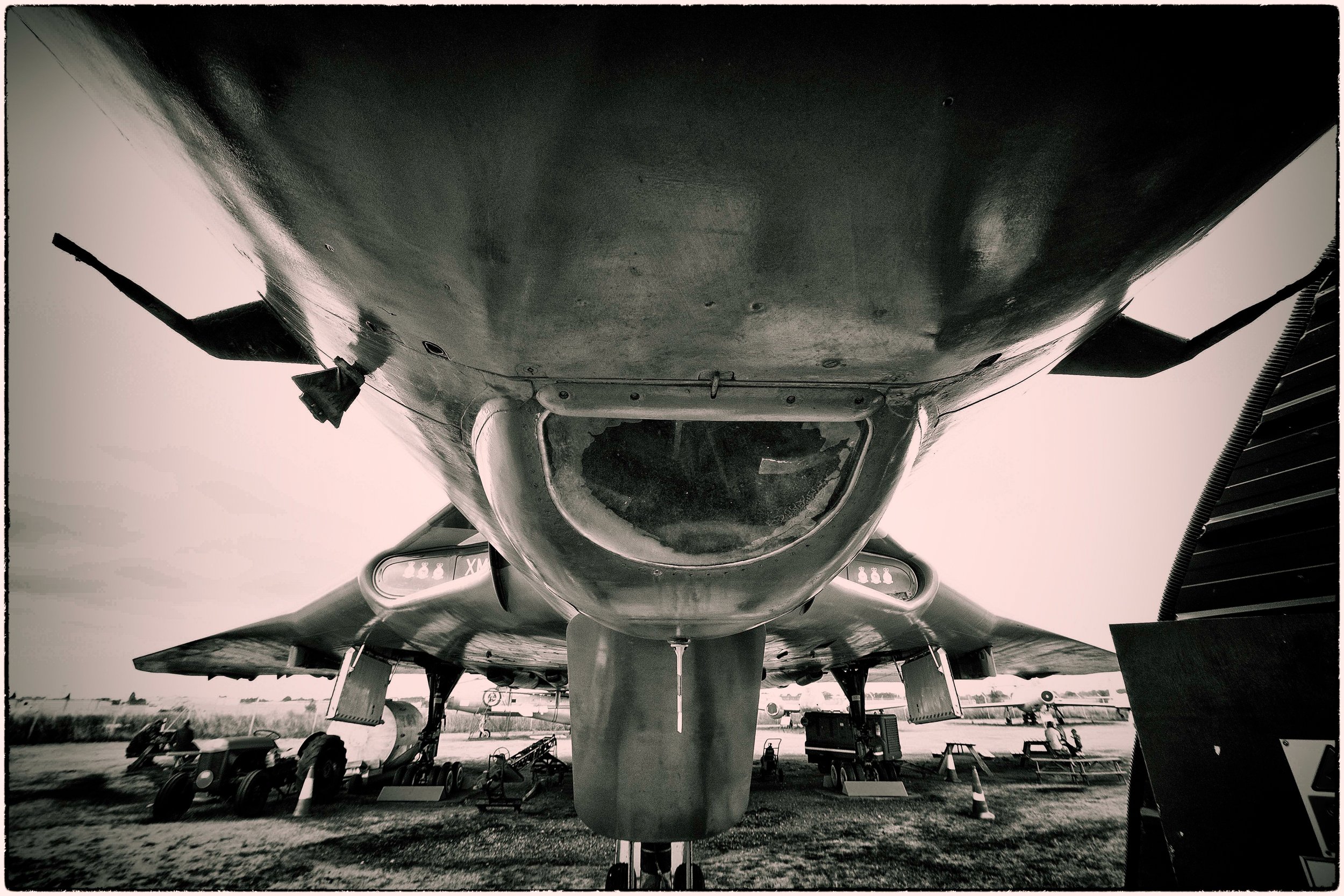 Black and white photo of the underside of a military jet aircraft parked outdoors with surrounding equipment and vehicles.