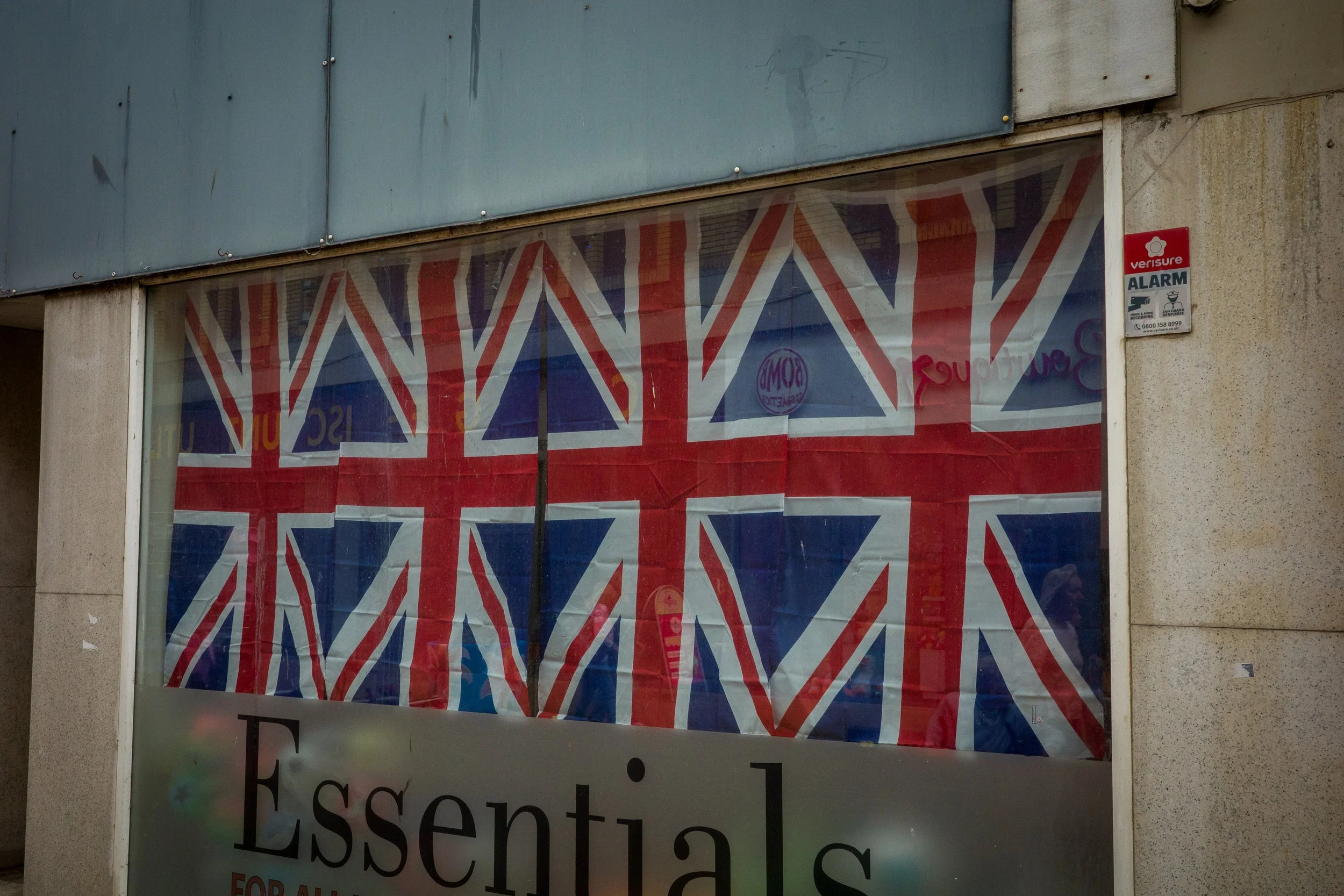 Storefront window with a Union Jack flag design and a security alarm sticker on the right side, part of a sign at the bottom is visible with the text starting 'Essen...'.