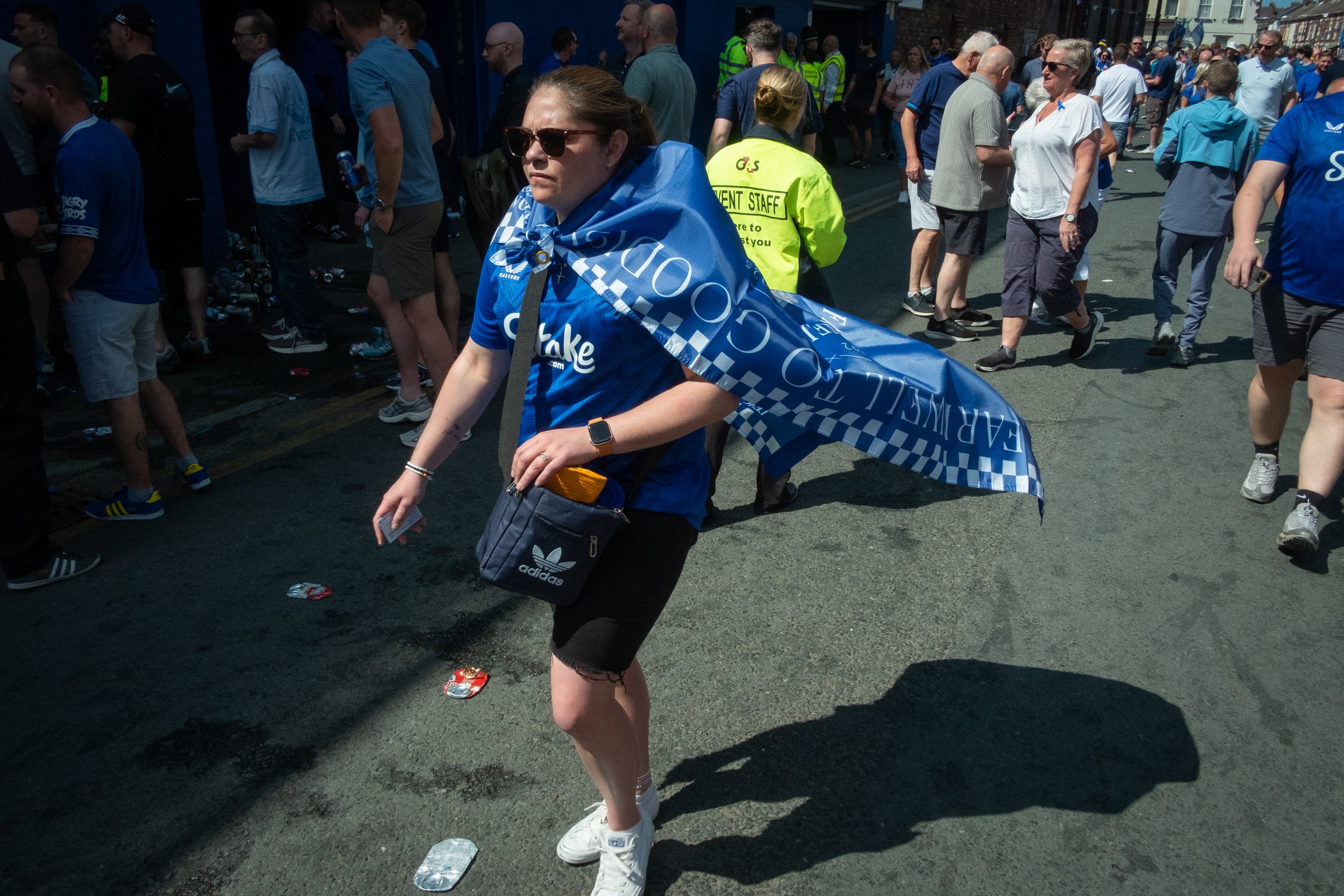A woman wearing sunglasses, a blue shirt, and a small black Adidas bag is walking through a crowded street after a sporting event, with discarded cans and trash on the ground.