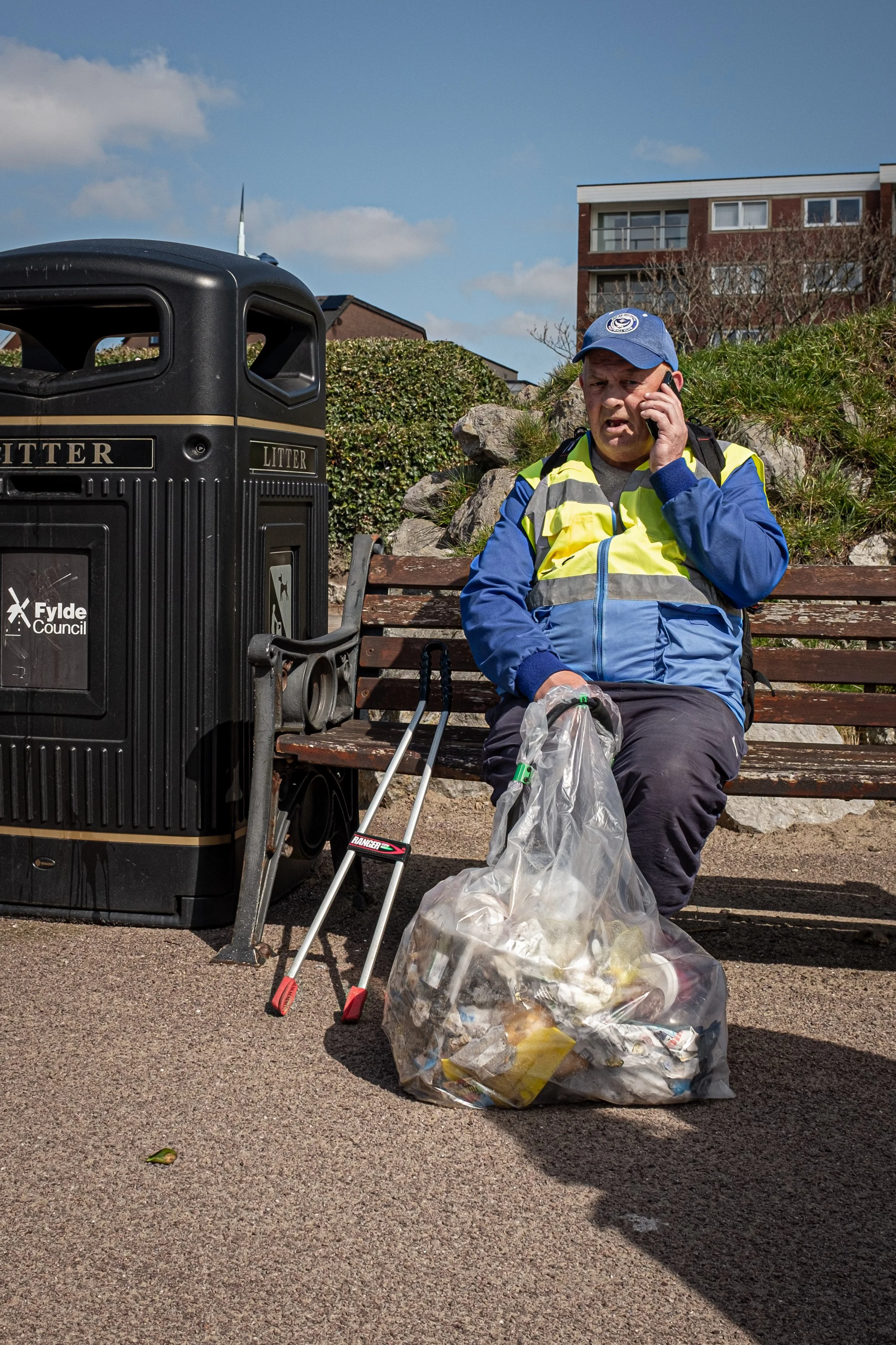 A man sitting on a park bench, talking on a cell phone, with trash bags and walking aid nearby, near a litter bin with 'Flyde Council' written on it, and residential buildings in the background.