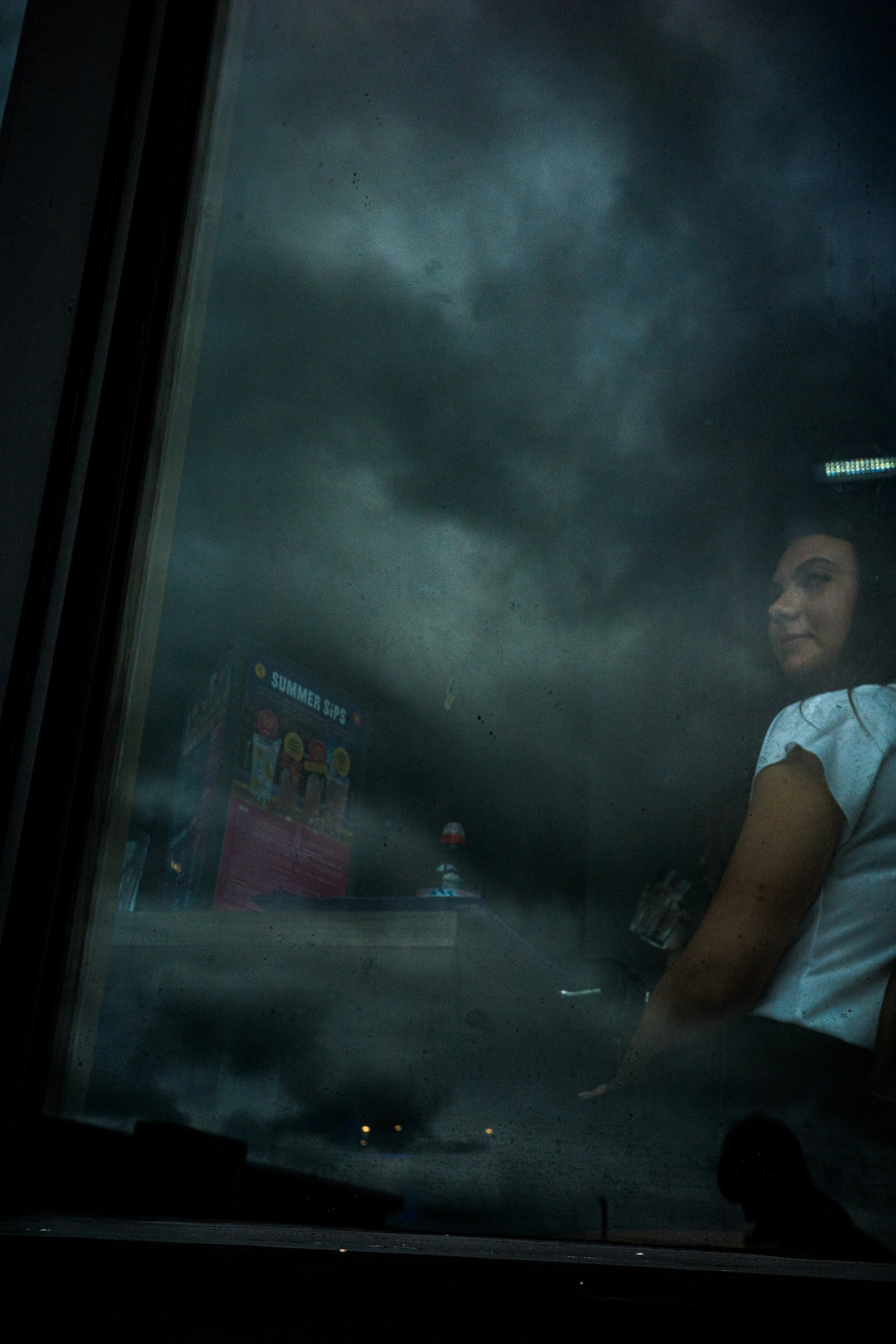Reflection of a woman with dark hair in a rainy window, with dark storm clouds and a glimpse of a vending machine and a person’s silhouette in the background.