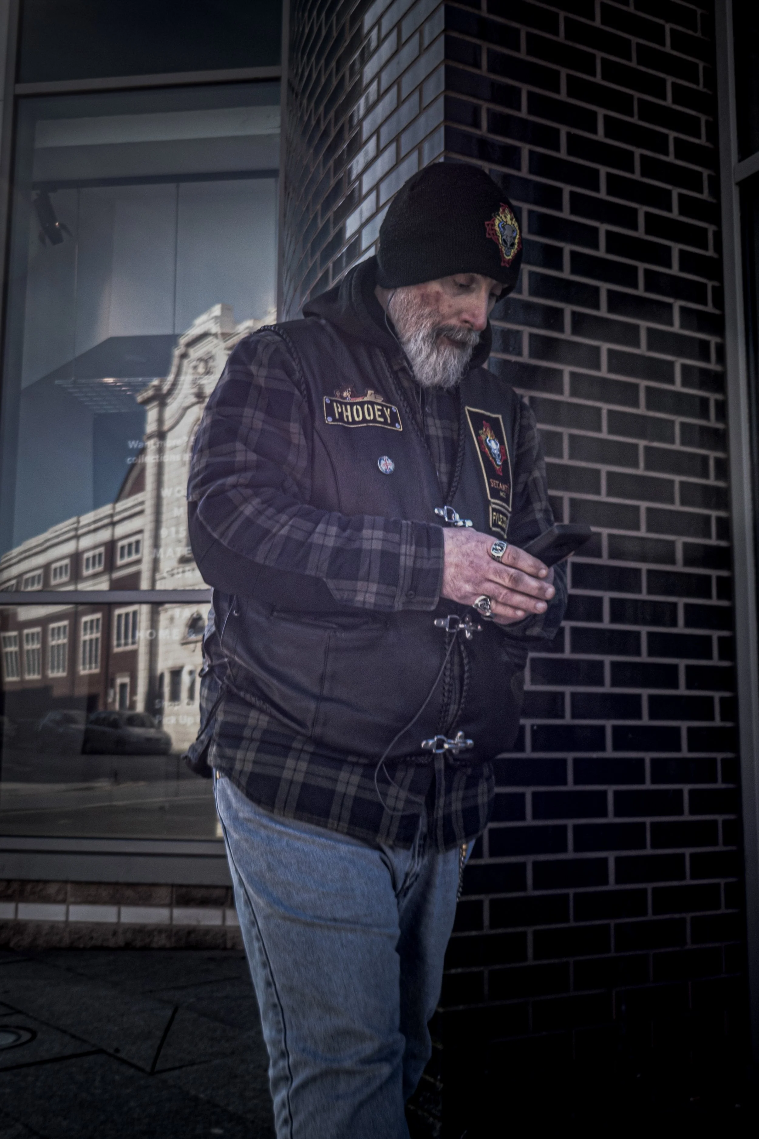 A man with a gray beard wearing a black beanie with a colorful emblem, a black vest with patches and a plaid shirt underneath, is looking at his phone while standing outdoors against a brick building and large glass windows.
