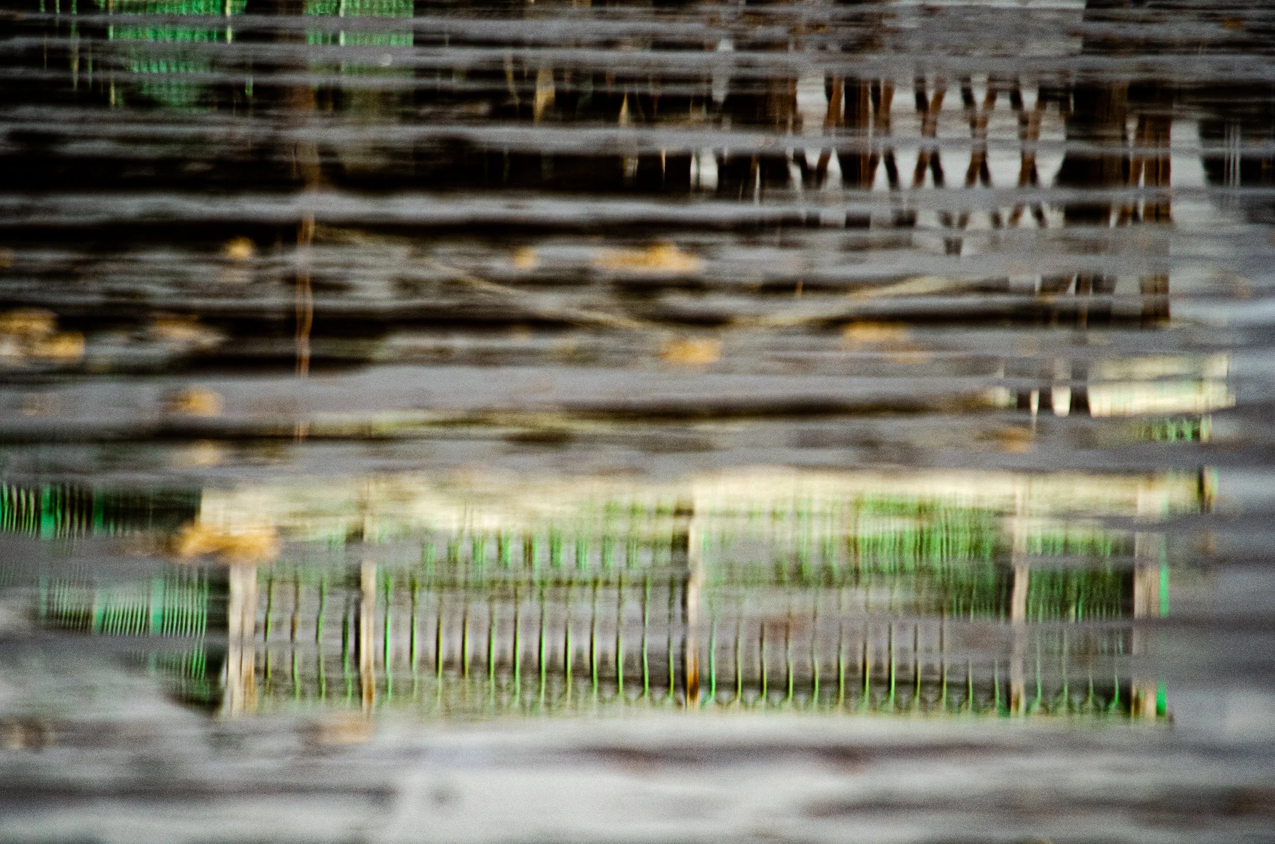 Reflection of a wooden and green outdoor structure in water, with ripples and distortions.