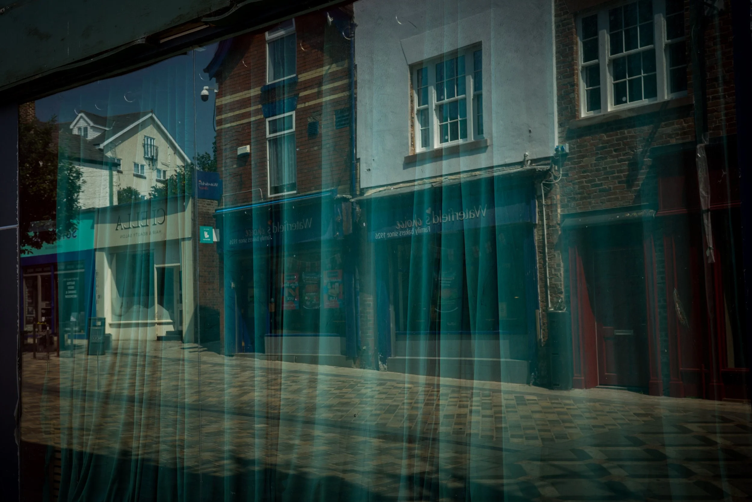 Reflected outside buildings with brick and painted exteriors; window with multiple panes; sidewalk with pattern; store sign inside store window reading 'Chelsea'.
