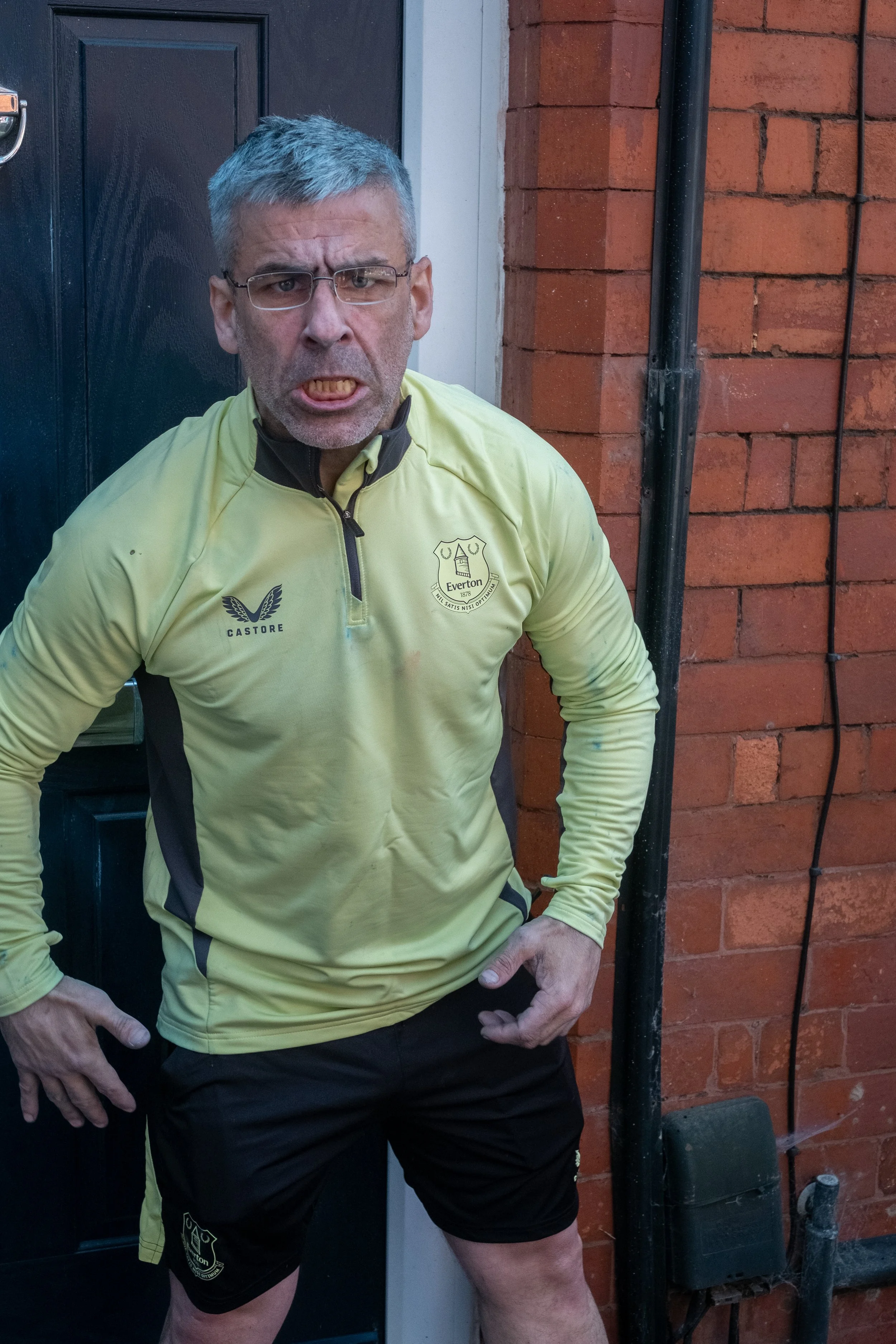 A man with gray hair and glasses, making an angry face, stands outside near a brick wall and a door, wearing a yellow sports jacket with the Everton football club logo.