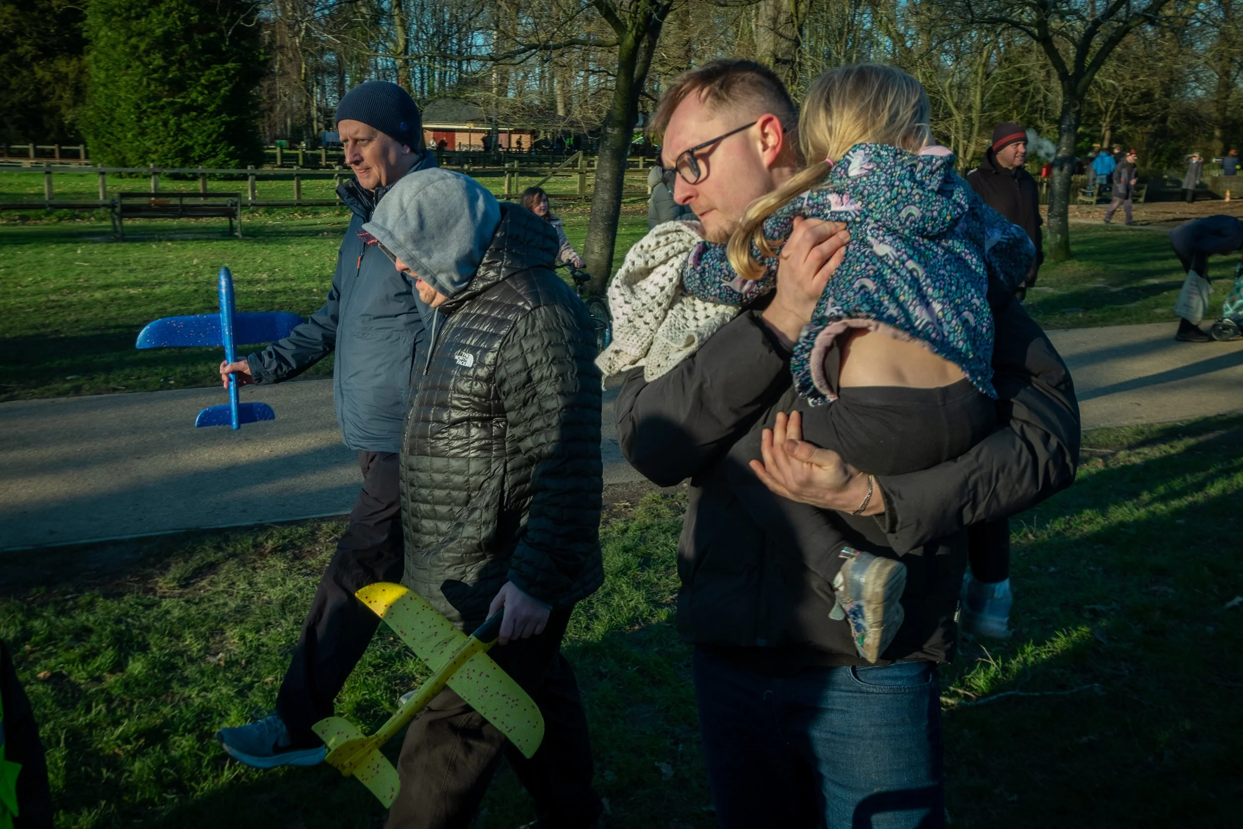 A family walking outdoors in a park on a sunny day, with a man carrying a young girl on his shoulders, and children playing nearby.