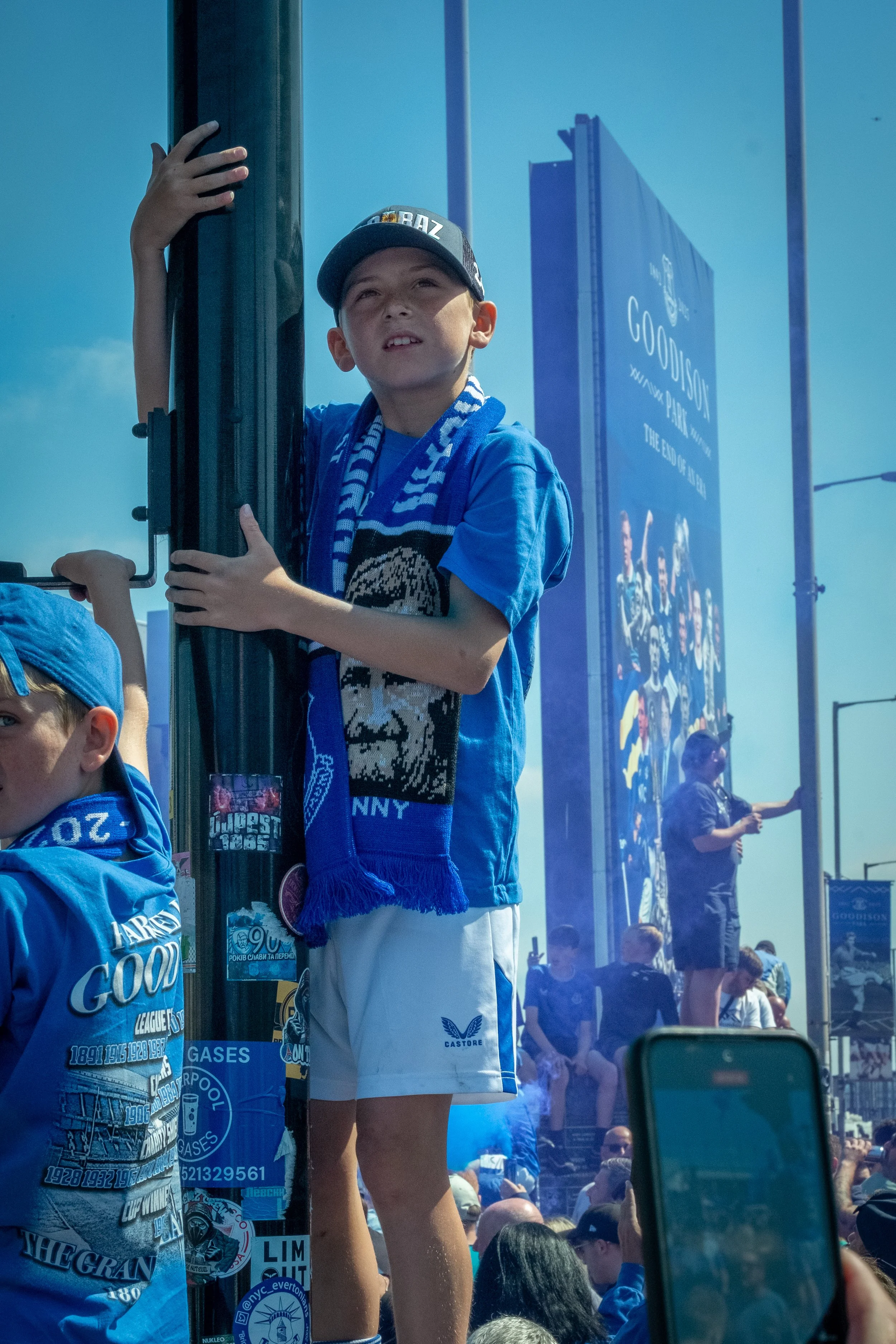 Young boy standing on a platform during a sports event, wearing a blue team scarf and cap, holding onto a pole, with a crowd and large billboard in the background.