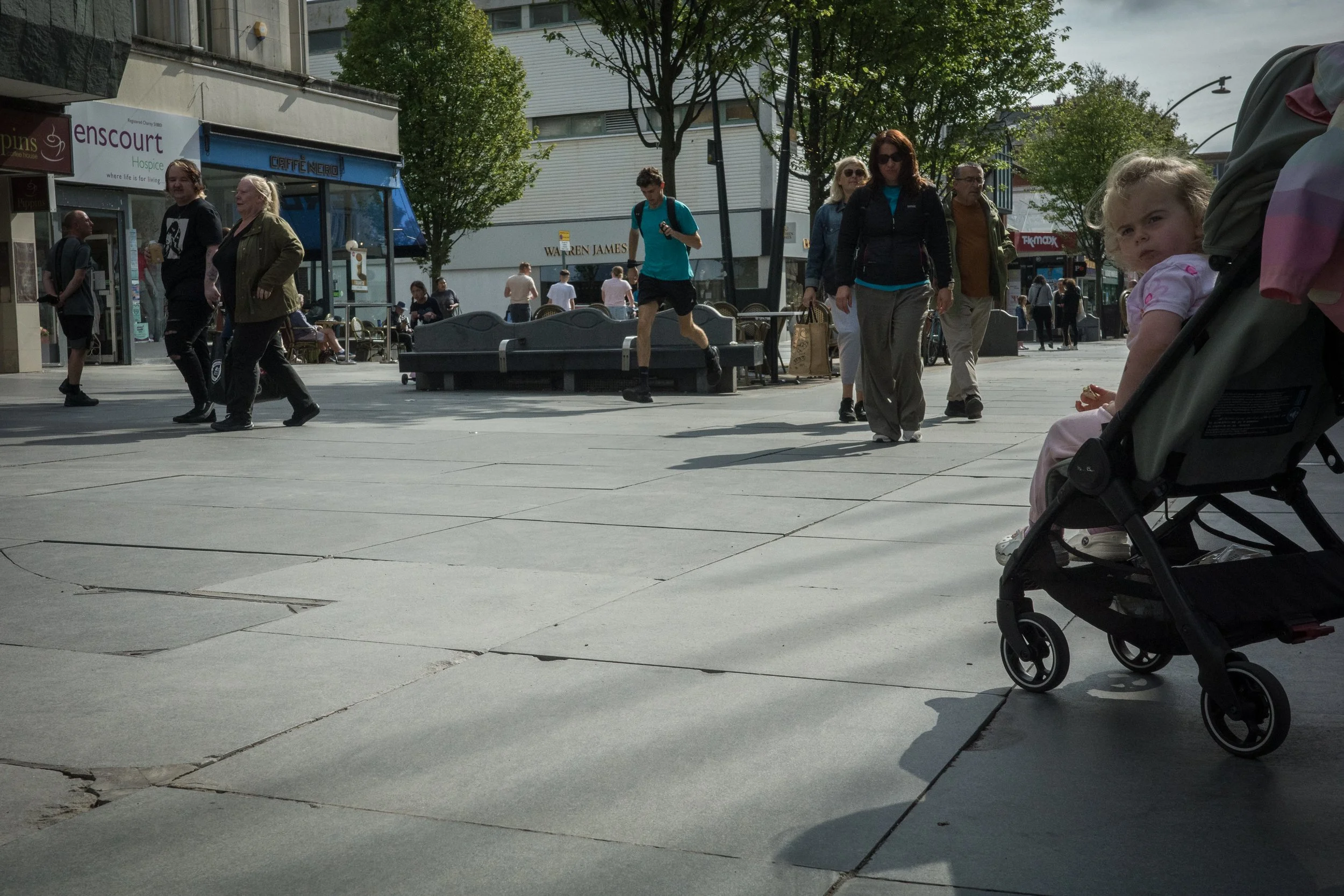 People walking on a city sidewalk with storefronts, trees, and a cloudy sky in the background.