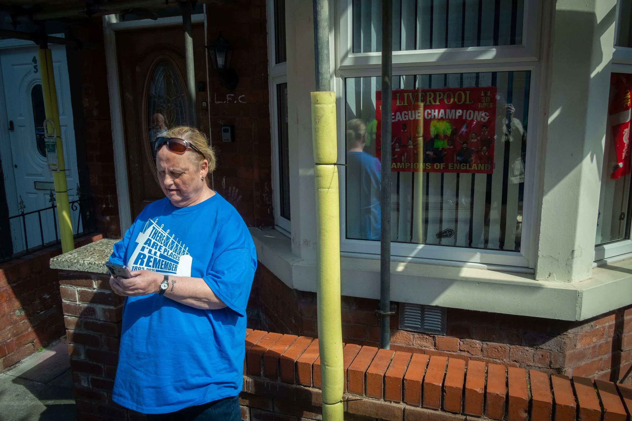 A woman in a blue shirt standing outside a house, looking at her phone, with a Liverpool Champions banner visible through the window behind her.