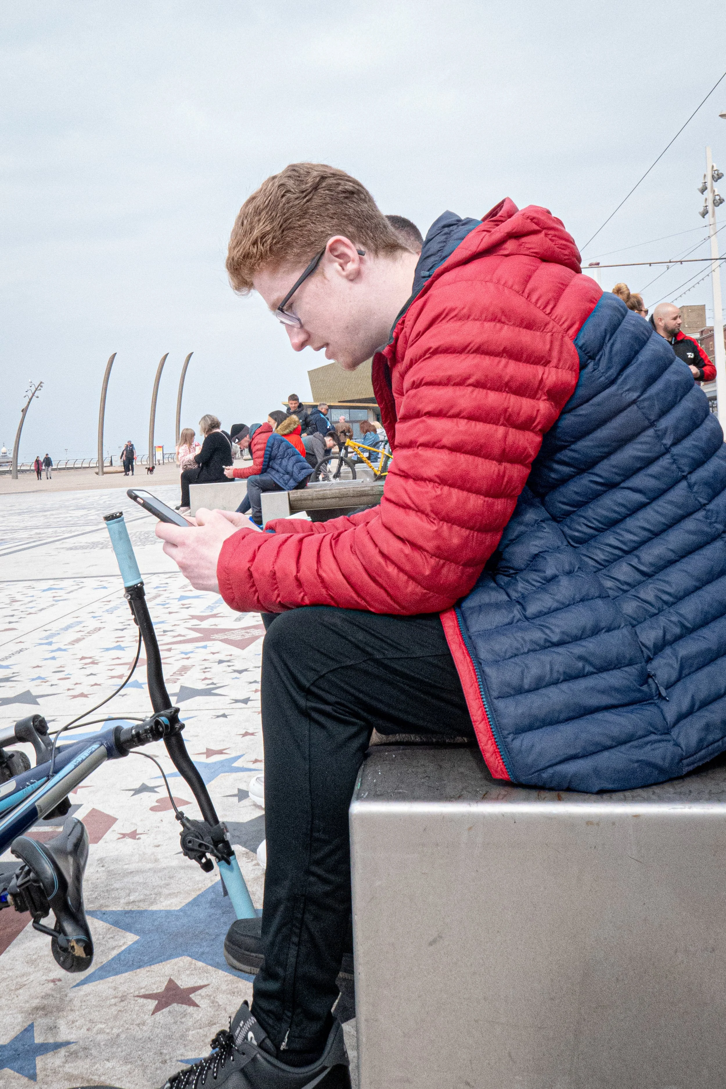 A young man with red hair and glasses wearing a red and blue puffy jacket, sitting on a bench on a city street, looking at his phone. A bicycle is visible in front of him, with stars on the sidewalk. People are sitting and walking in the background n