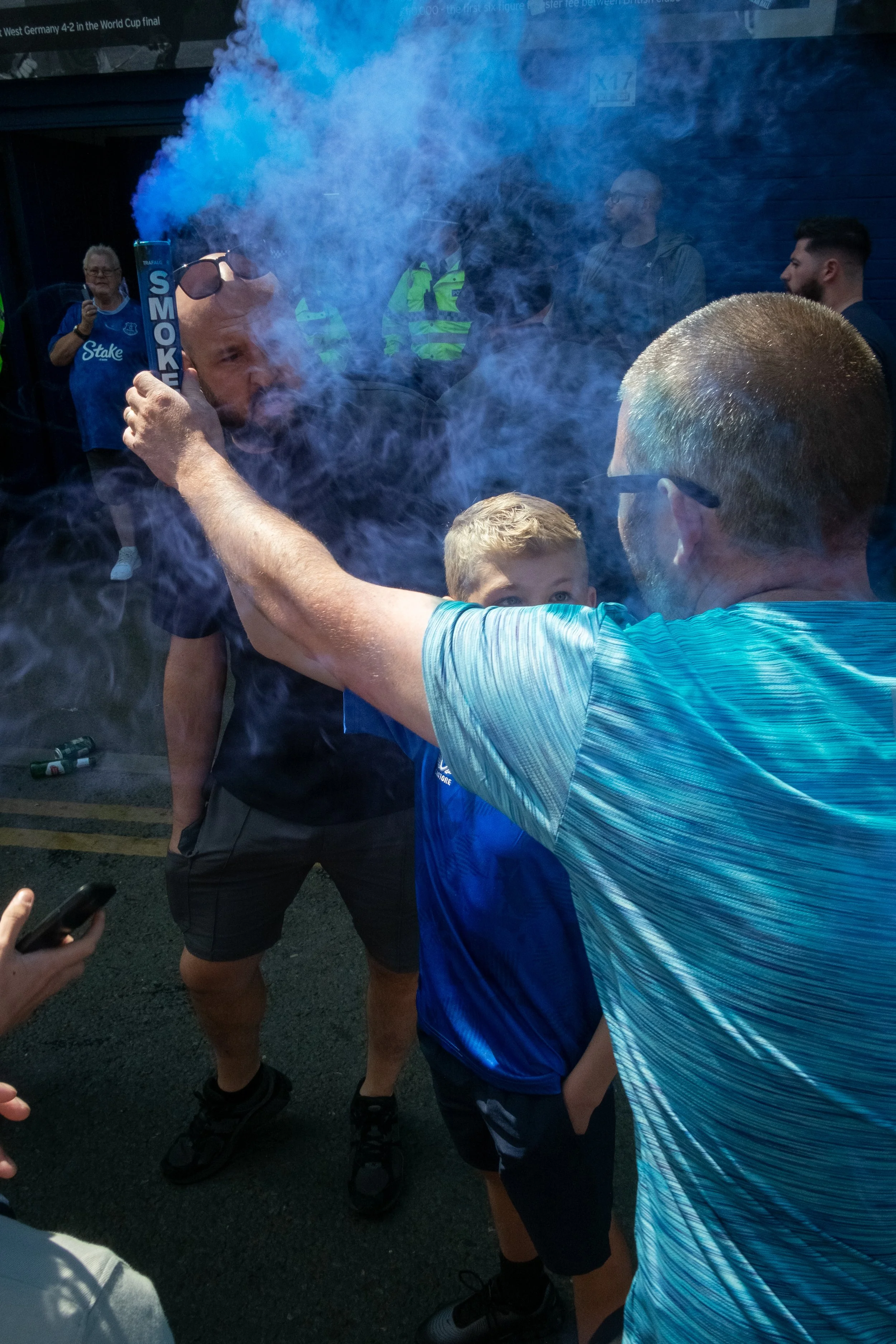 A man in a blue shirt holds a smoke canister, releasing blue smoke, while a young boy in a blue shirt looks up at him amid a crowd.