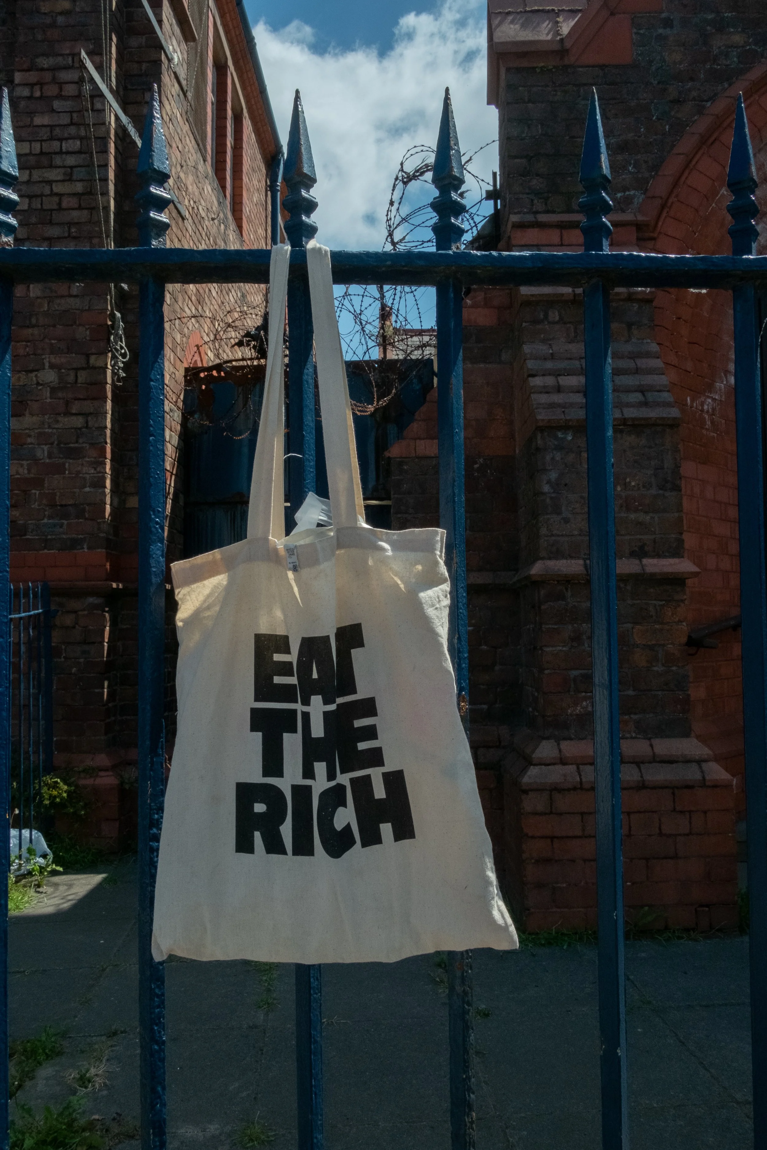 A fabric tote bag hanging on a blue metal fence, with a brick building in the background and the sky visible above