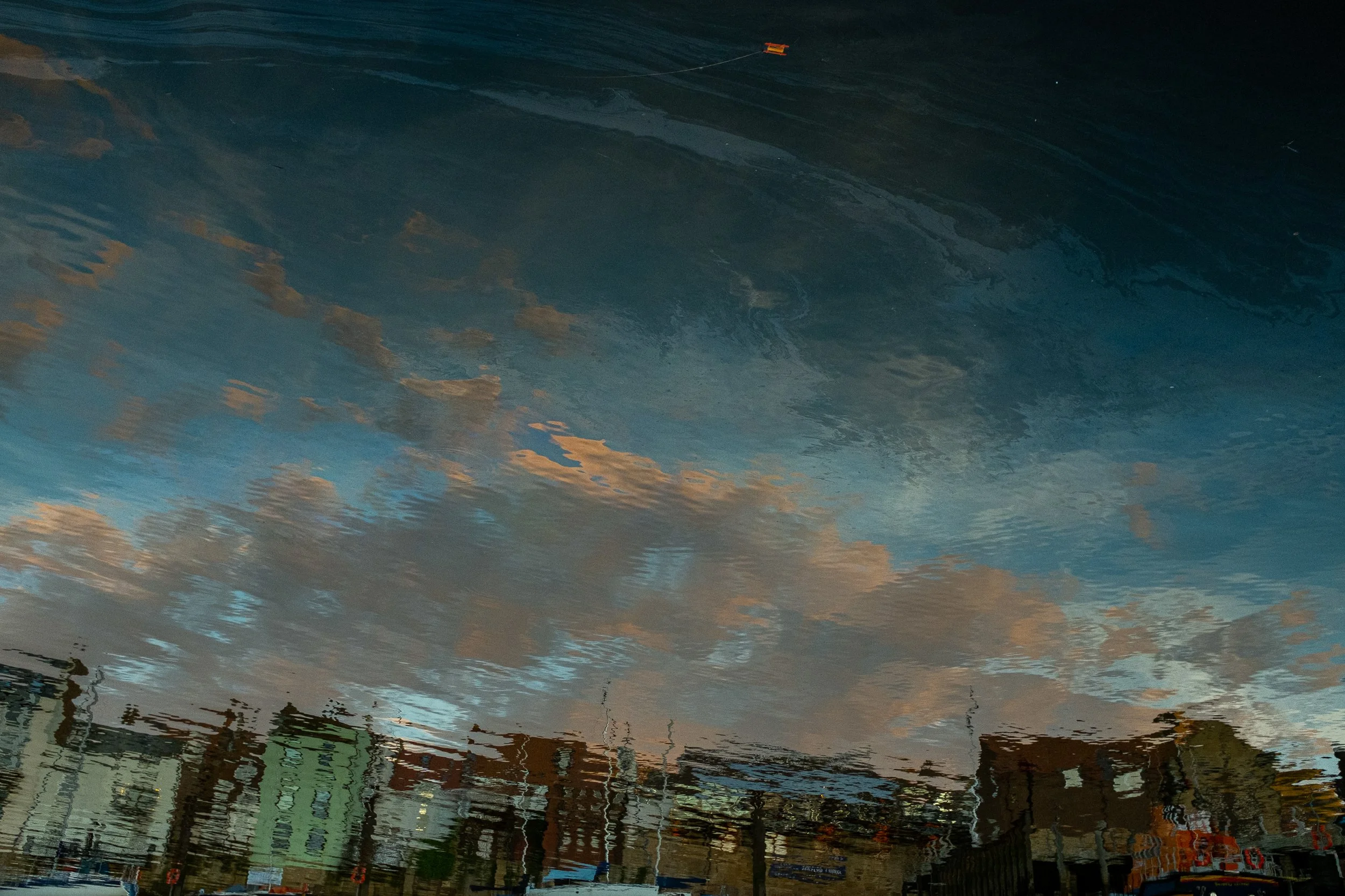 Reflections of a cloudy sky and buildings in a puddle on the ground.