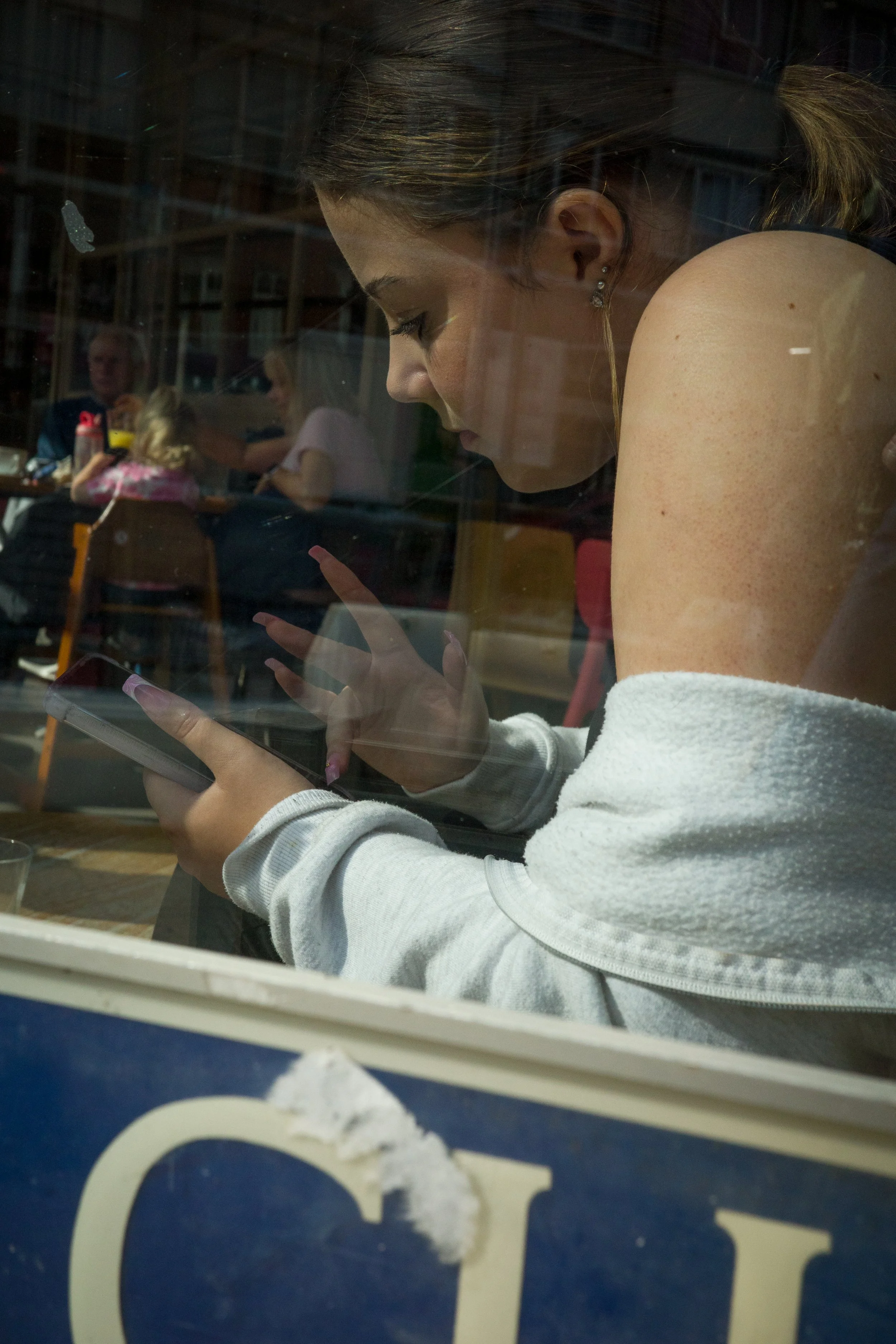 Woman with brown hair looking at her phone, seen through a window with reflections of people sitting at tables inside a cafe or restaurant.