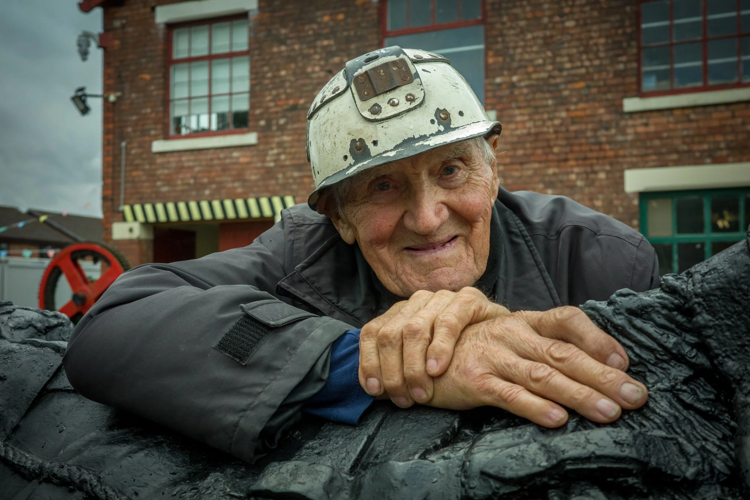 An elderly man wearing a white construction helmet, smiling, and resting his head on a textured surface outdoors near a brick building.