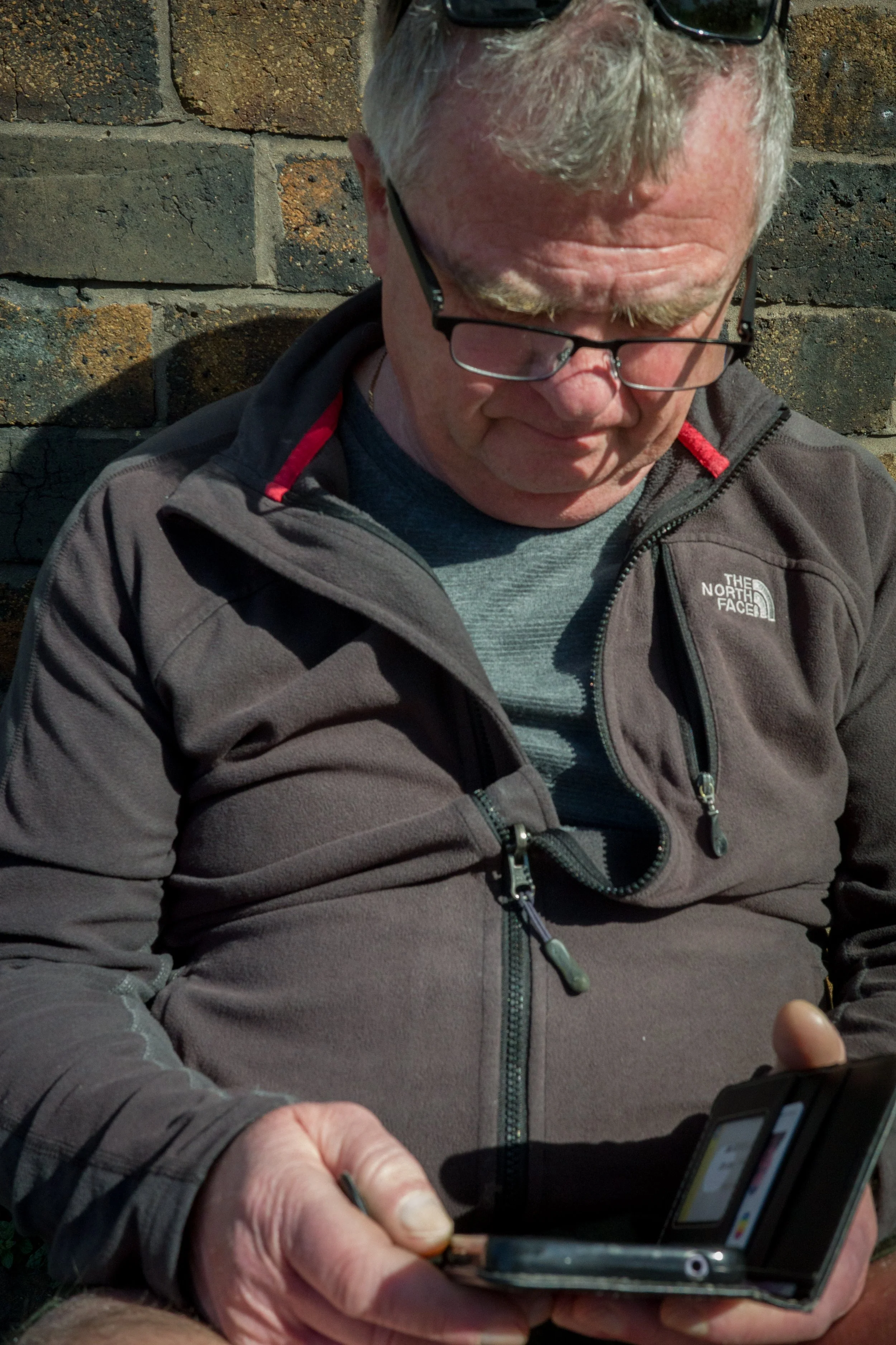 A man with gray hair and glasses wearing a dark gray North Face jacket, looking down at a mobile phone.