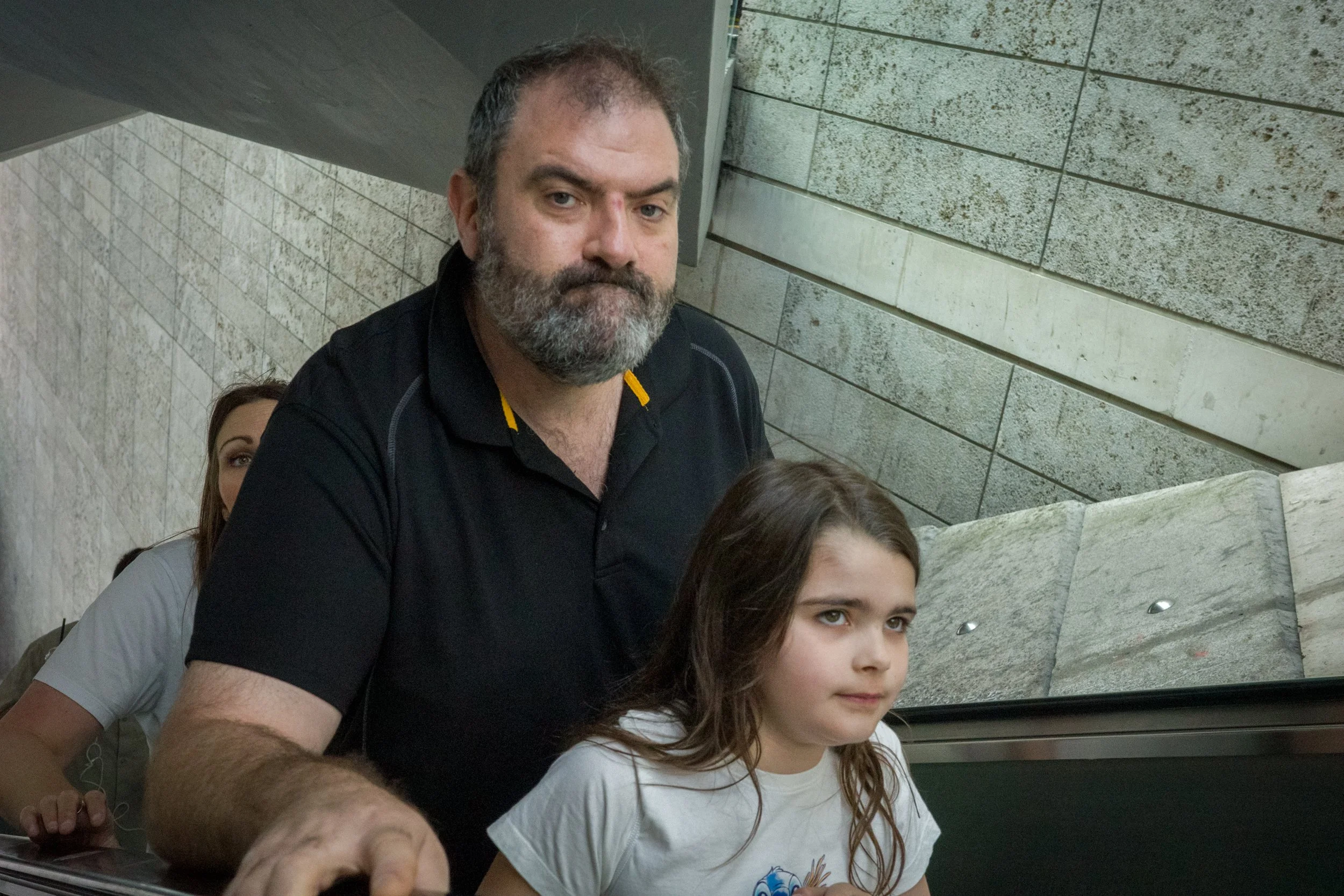 A bearded man and a young girl with brown hair are sitting on an escalator inside a building with stone wall panels.