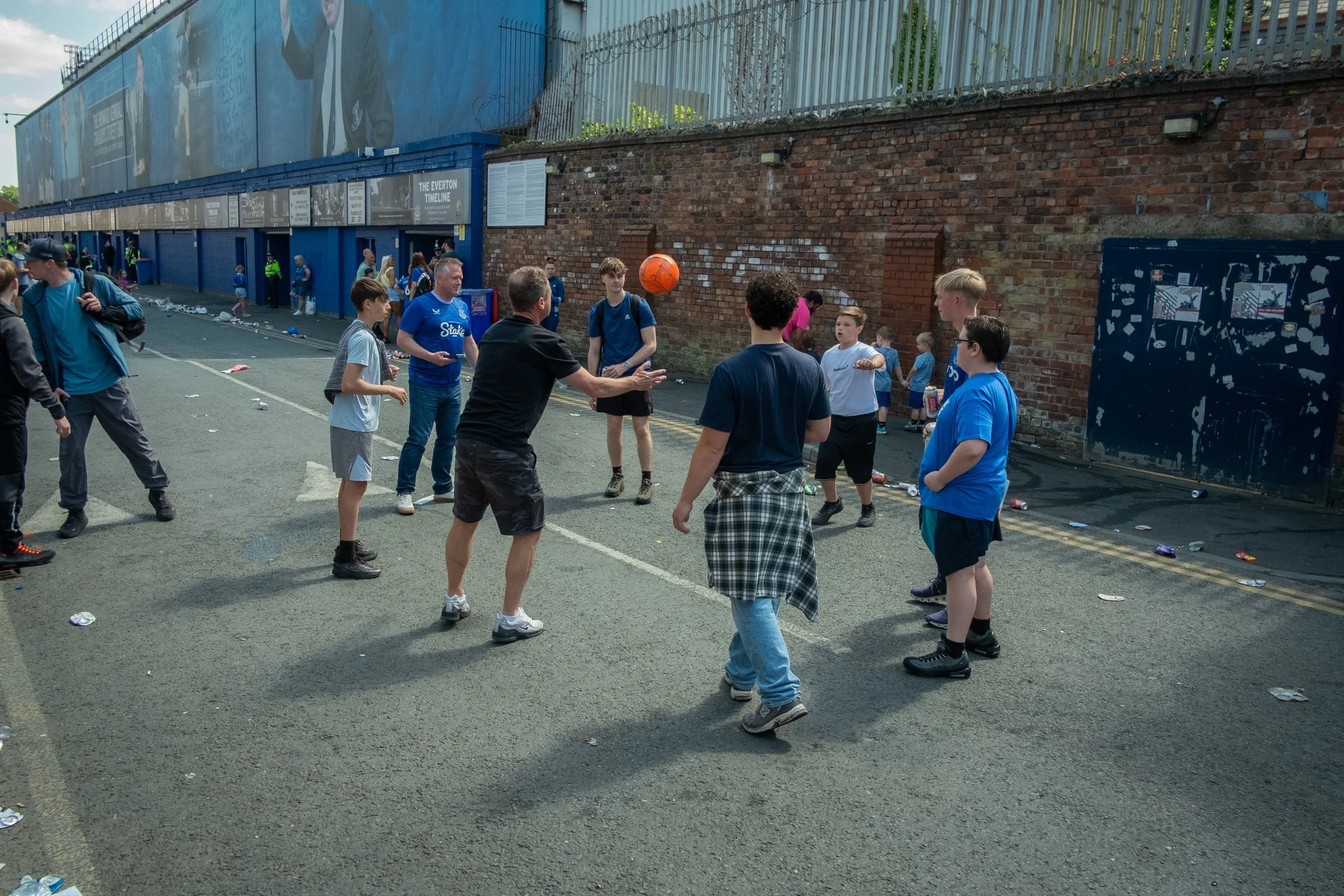 A group of young people and adults playing football on an urban street with a brick wall and a building in the background, littered with trash.