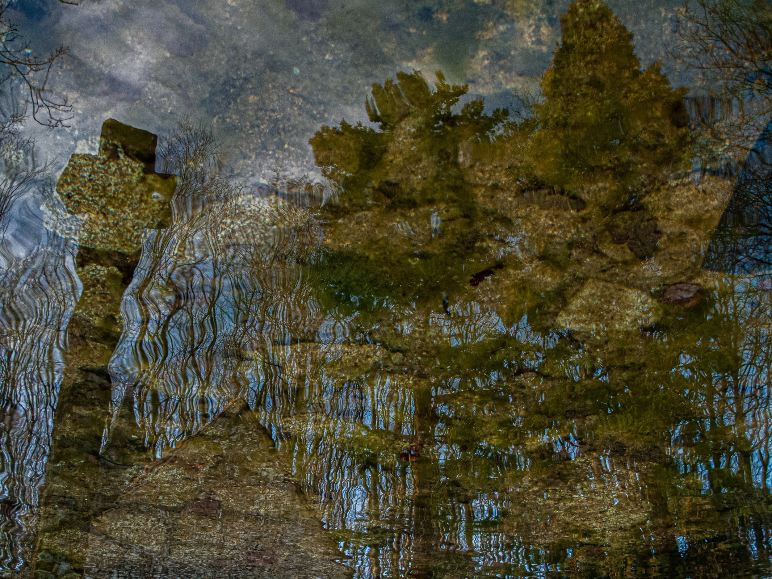 Reflection of trees on a shallow, rocky stream, with ripples in the water.