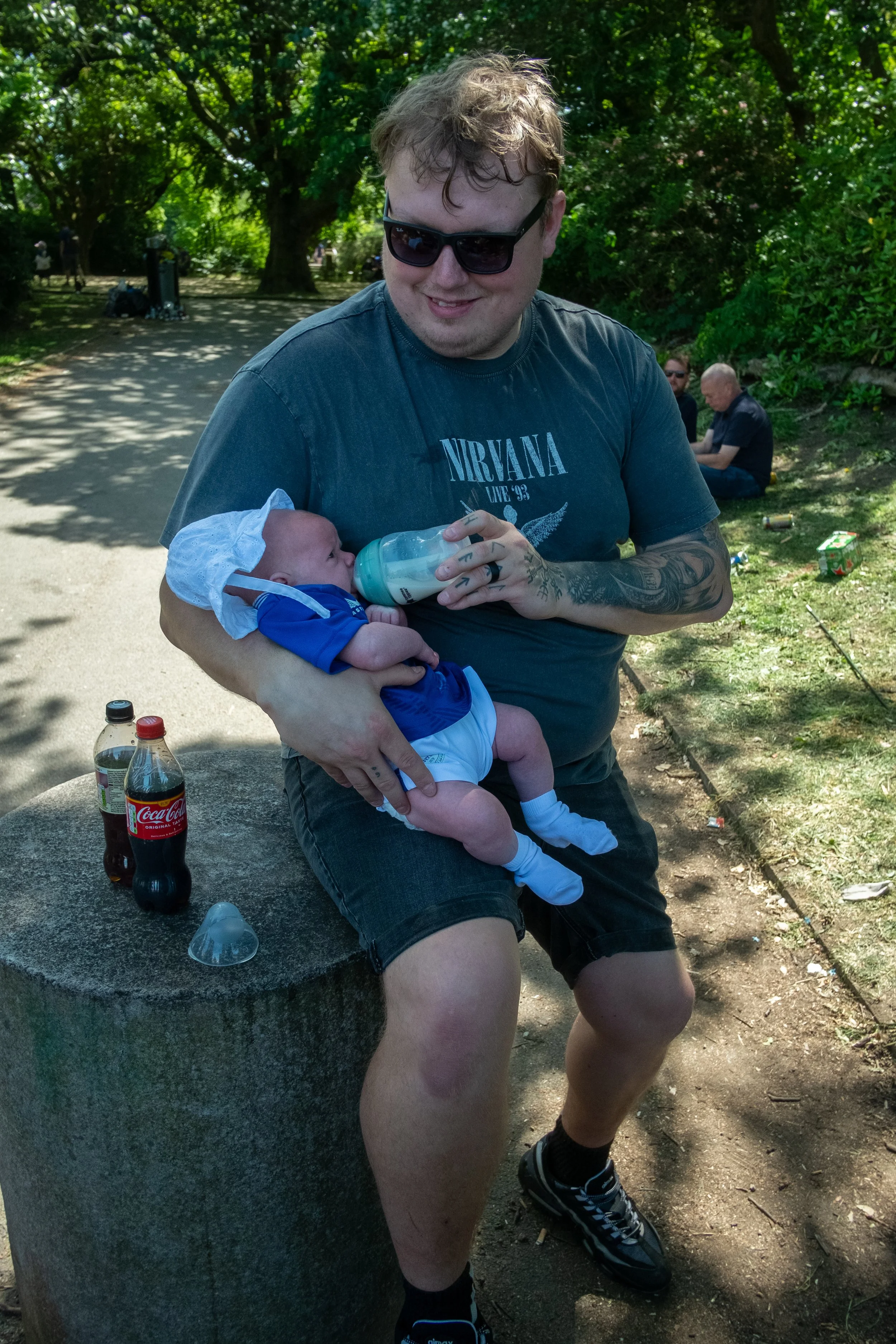 A man wearing sunglasses and a Nirvana T-shirt sitting on a stone bench in a park, feeding a baby with a bottle. The baby is dressed in a blue outfit with a white hat and socks, and is sitting on the man's lap. Two soda bottles and a small container 