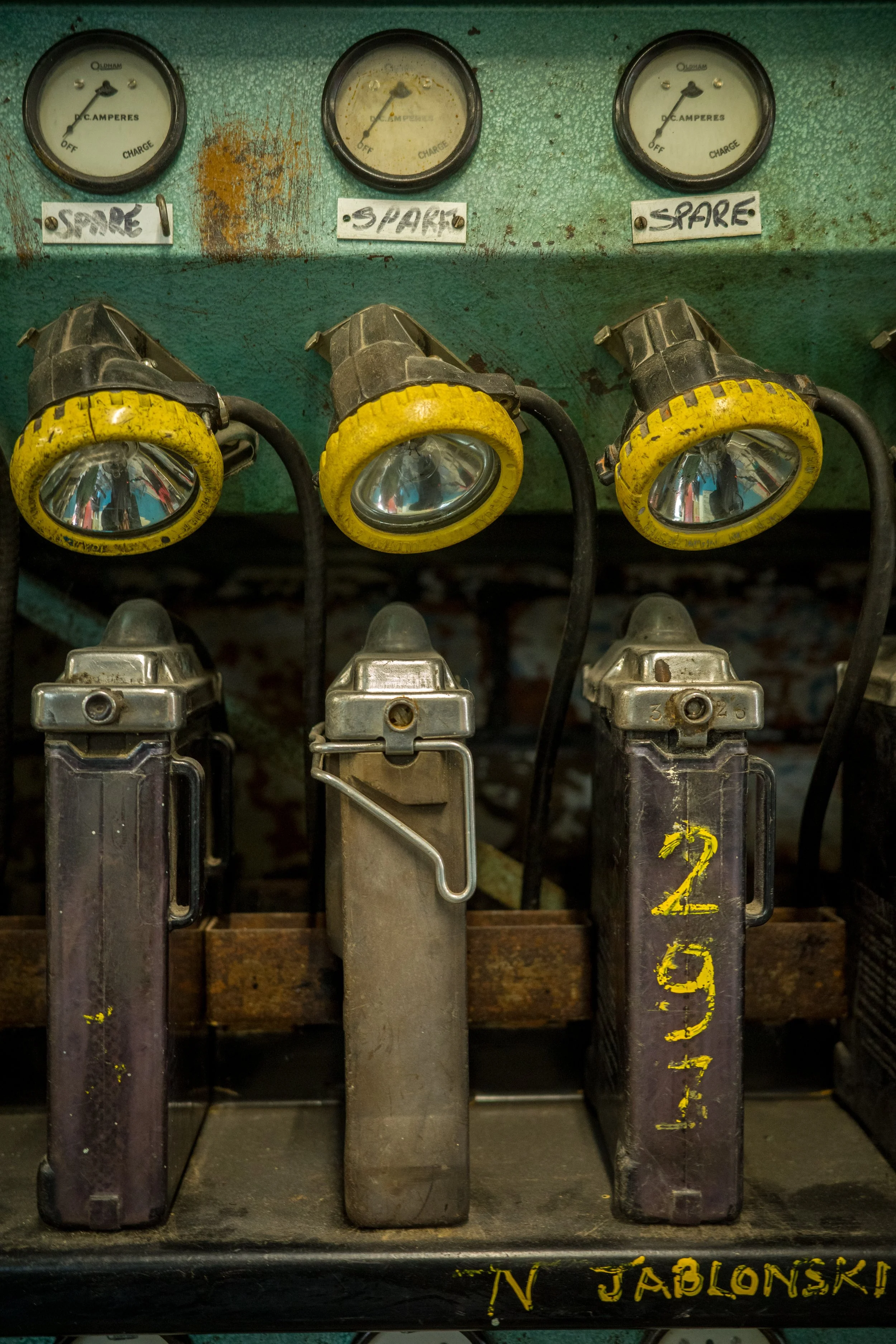 Four vintage machine guns with yellow and black labels, mounted on a green, rusted metal surface with gauge meters above them, indicating voltage or current, and handwritten labels below each gun reading 'SPARE', '3 SPARE', and another partially visi
