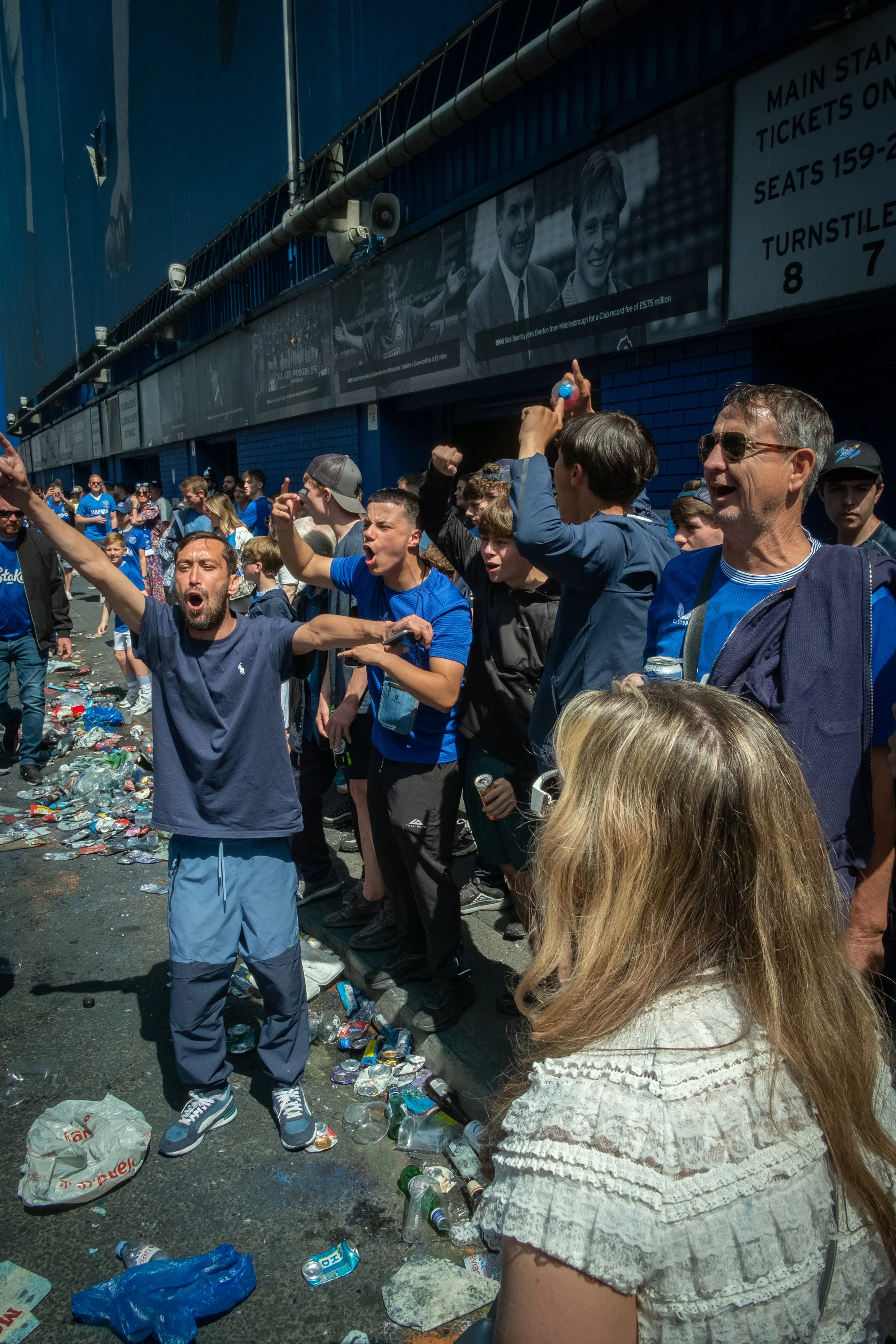 A group of people standing outside a stadium with trash scattered on the ground. They are celebrating, cheering, and some appear upset or frustrated.