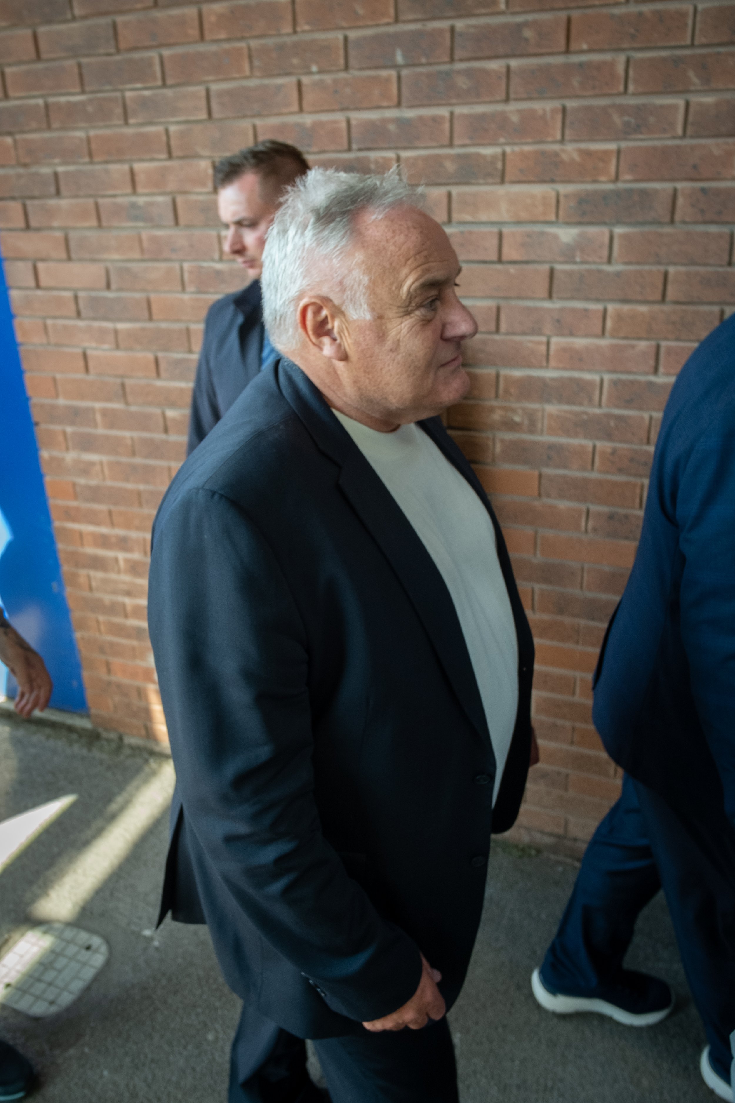 Older man with gray hair wearing a black blazer and white shirt standing against a brick wall.