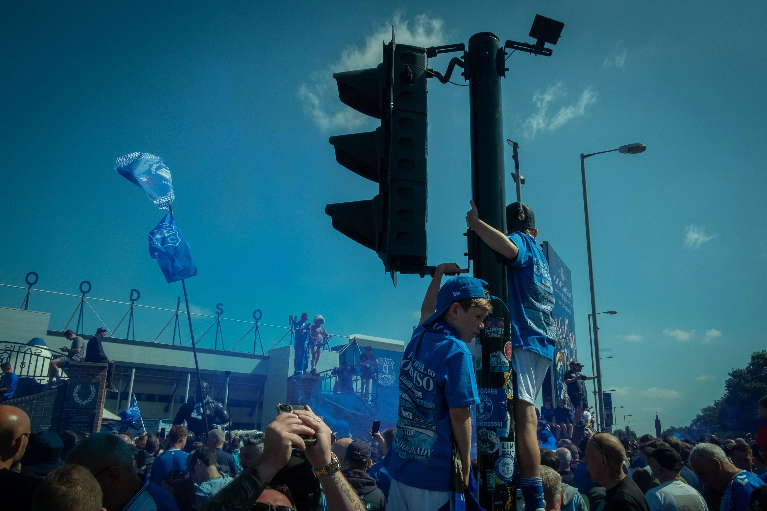 A large crowd gathered outdoors, some standing on a platform holding onto a pole and traffic lights, with people dressed in blue shirts. Flags and banners are visible, and a building in the background has the words 'CASH ODS' and 'GATES' on it.