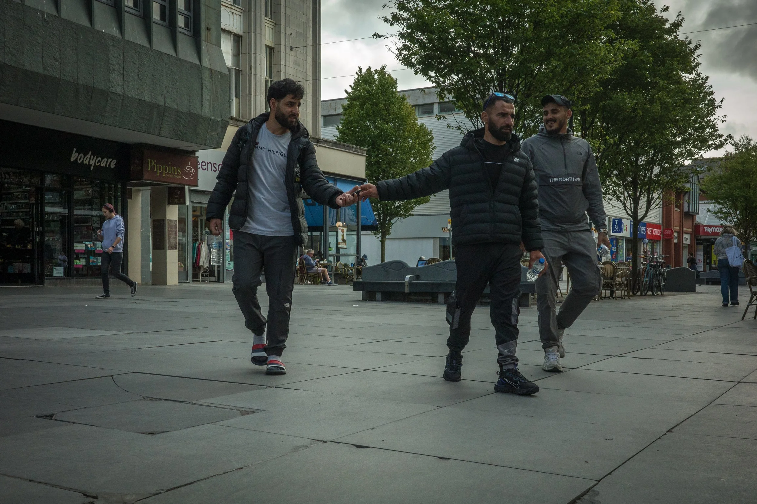 Three men walking on a city sidewalk, one extending his hand to shake hands with another, while the third man walks beside them.