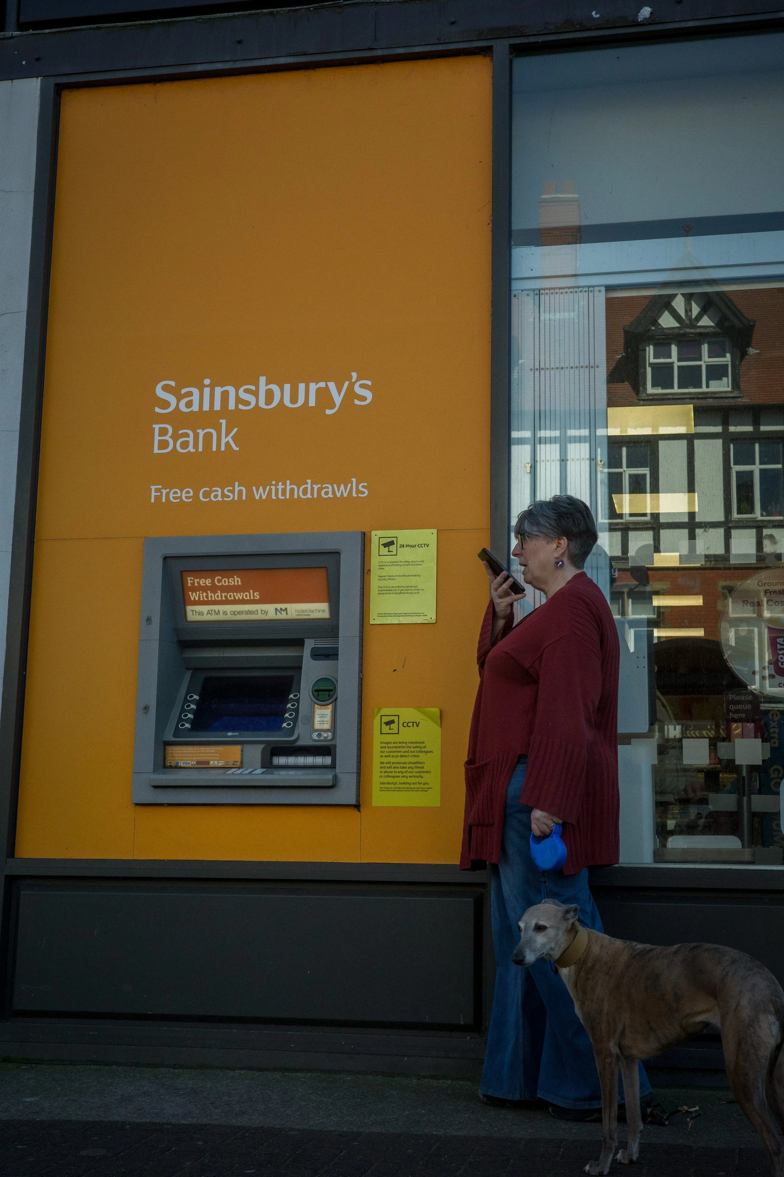 A woman wearing glasses and a red cardigan stands next to a large dog at a cash machine outside a Sainsbury's Bank branch. The woman is looking at her phone. The ATM offers free cash withdrawals and is operated by NatWest. There is a yellow sign indi