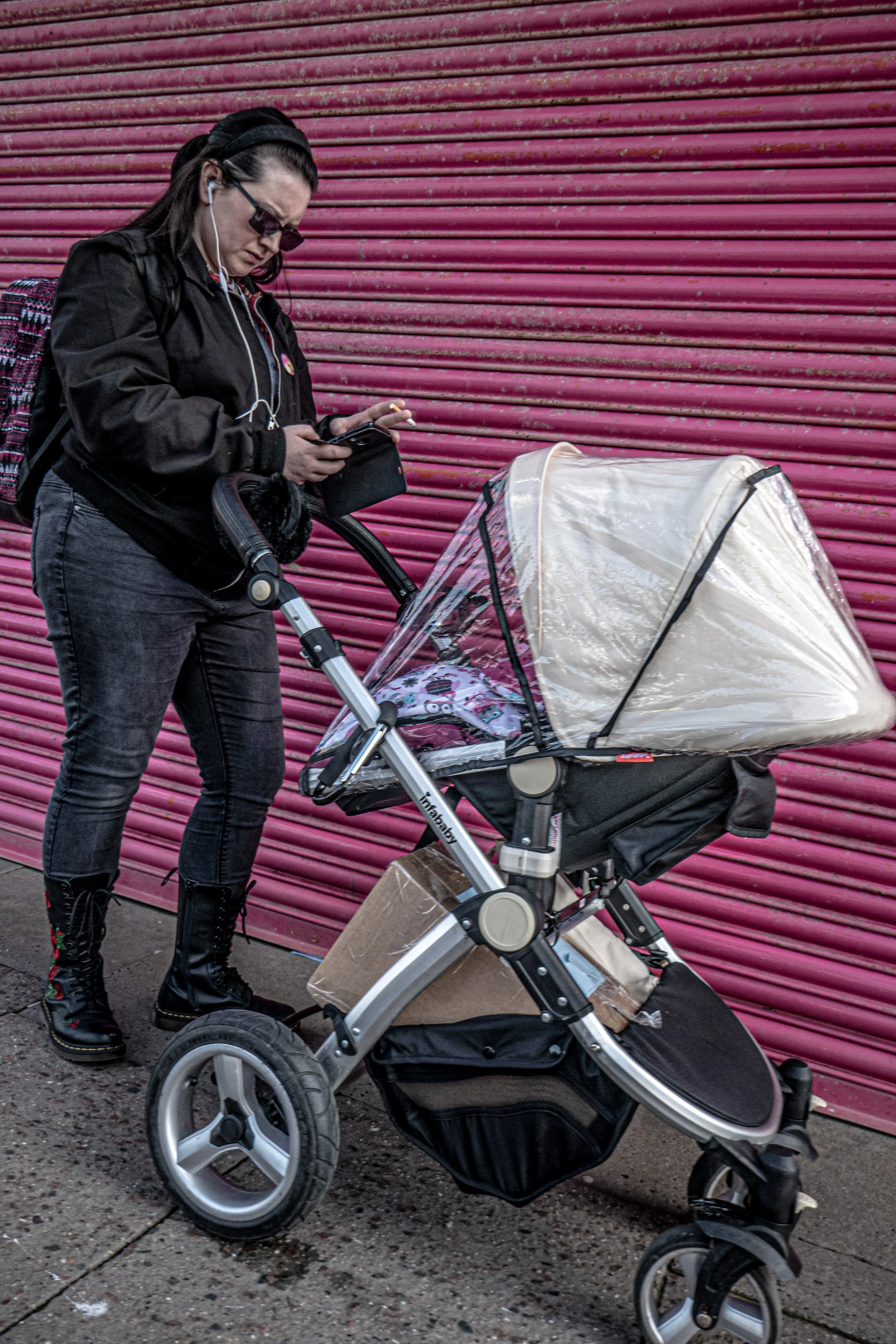 A woman wearing black clothing, sunglasses, and earphones, standing on a sidewalk, looking at her phone, next to a baby stroller with a transparent canopy, in front of a pink metal roll-up door.