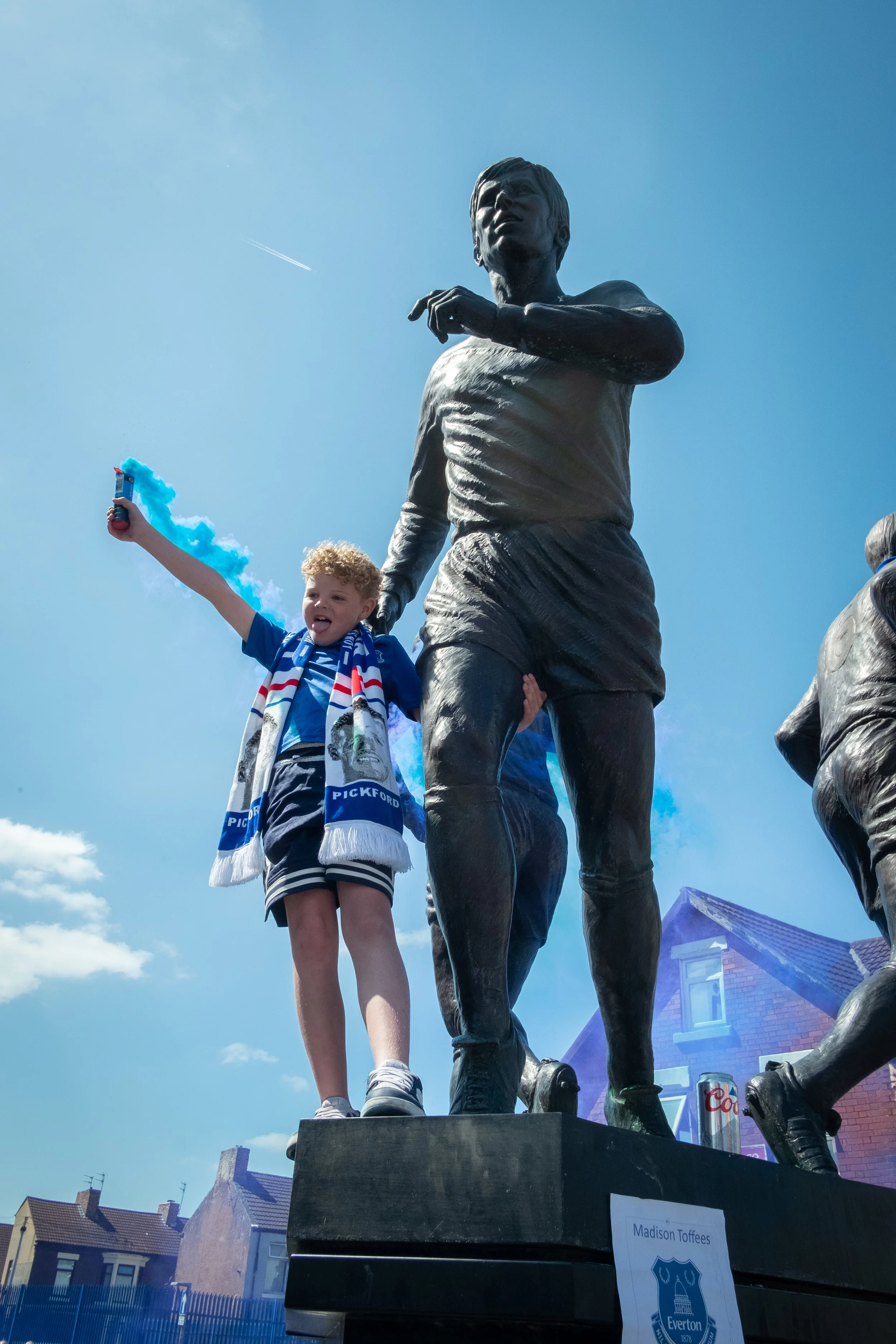 A young boy standing on a statue platform, holding a blue smoke flare, celebrating outside on a sunny day, with houses in the background.