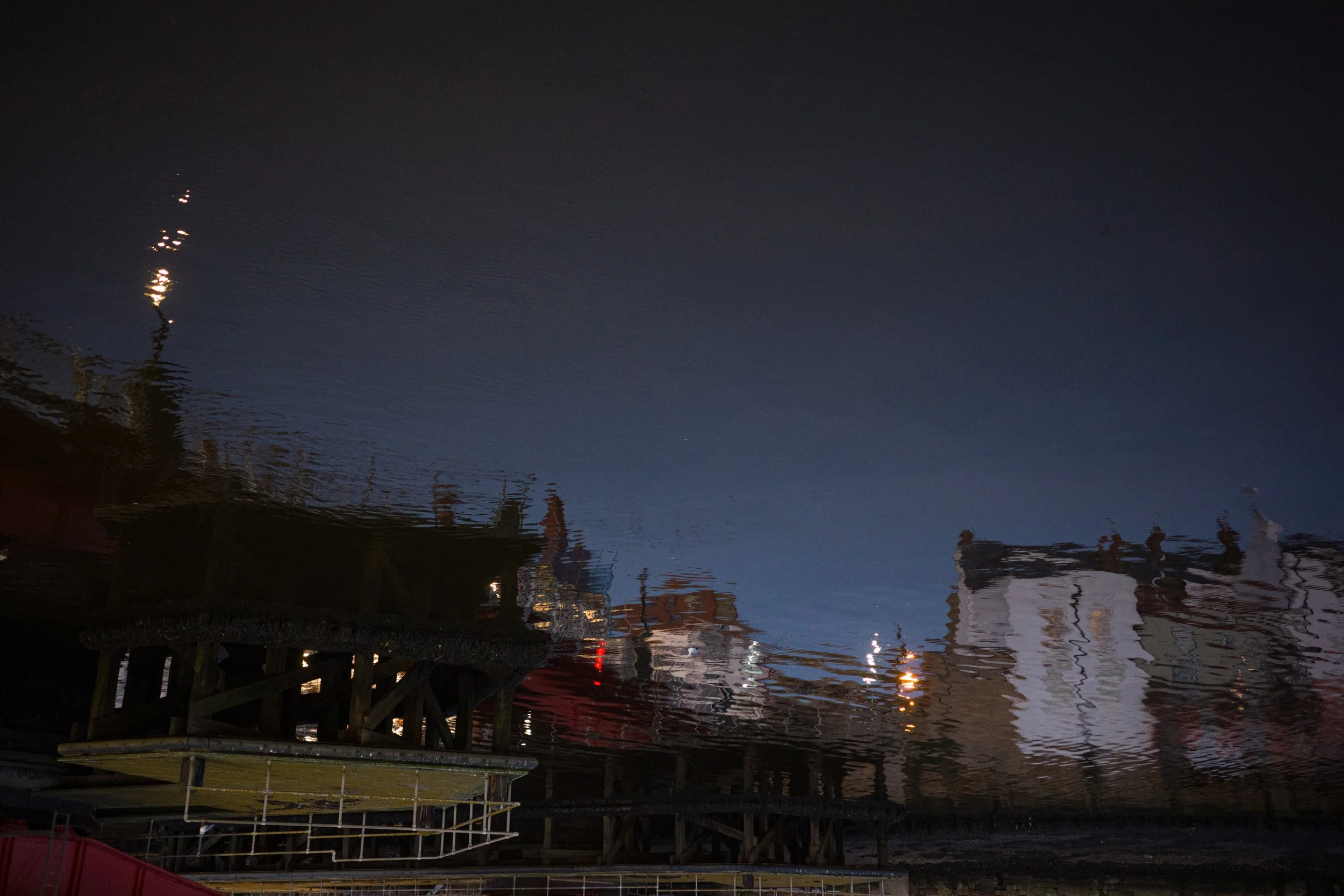 Reflection of buildings, streetlights, and a lighthouse in a body of water at night.